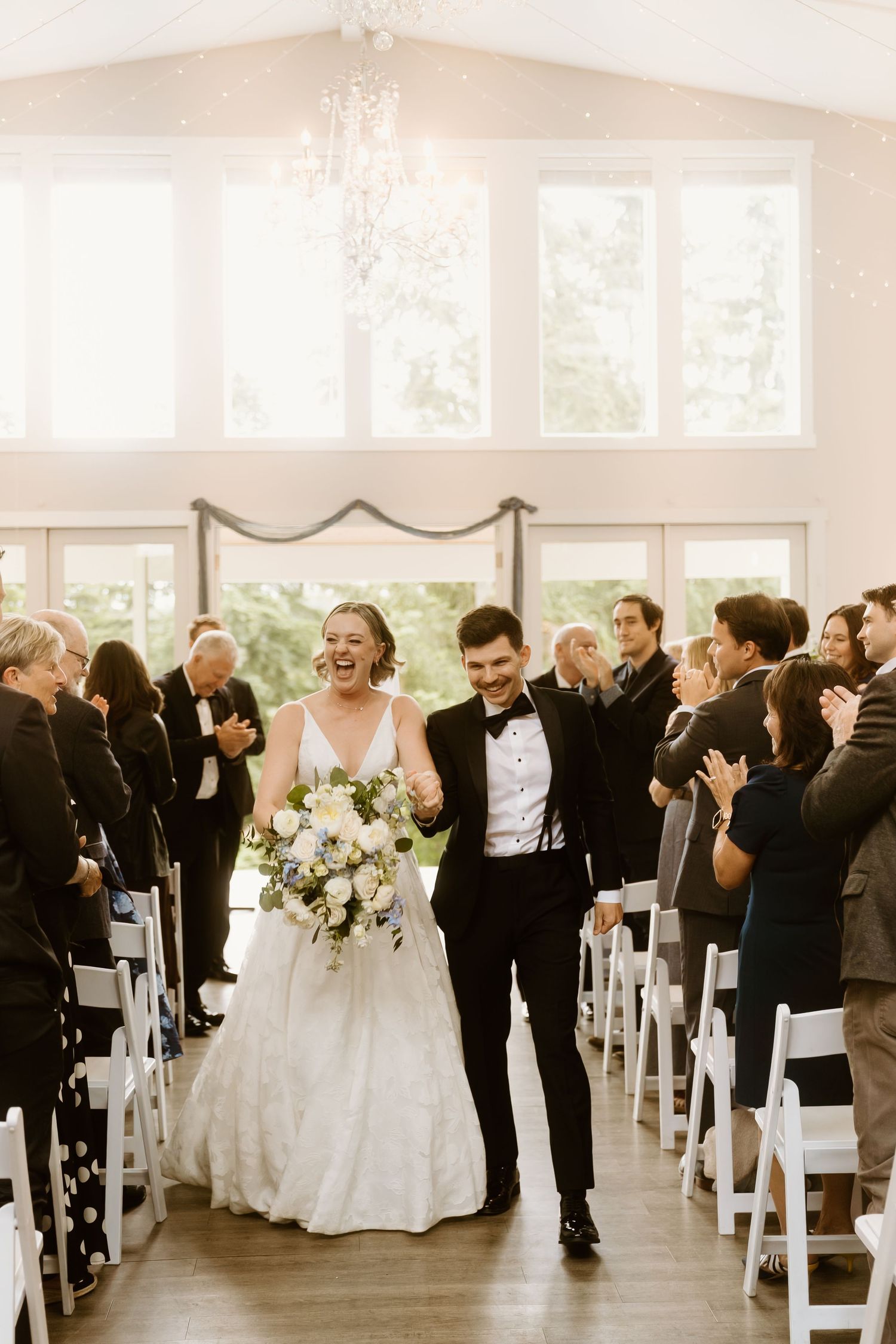 Joyful bride and groom walk down the aisle after wedding ceremony in elegant white chapel with chandelier.