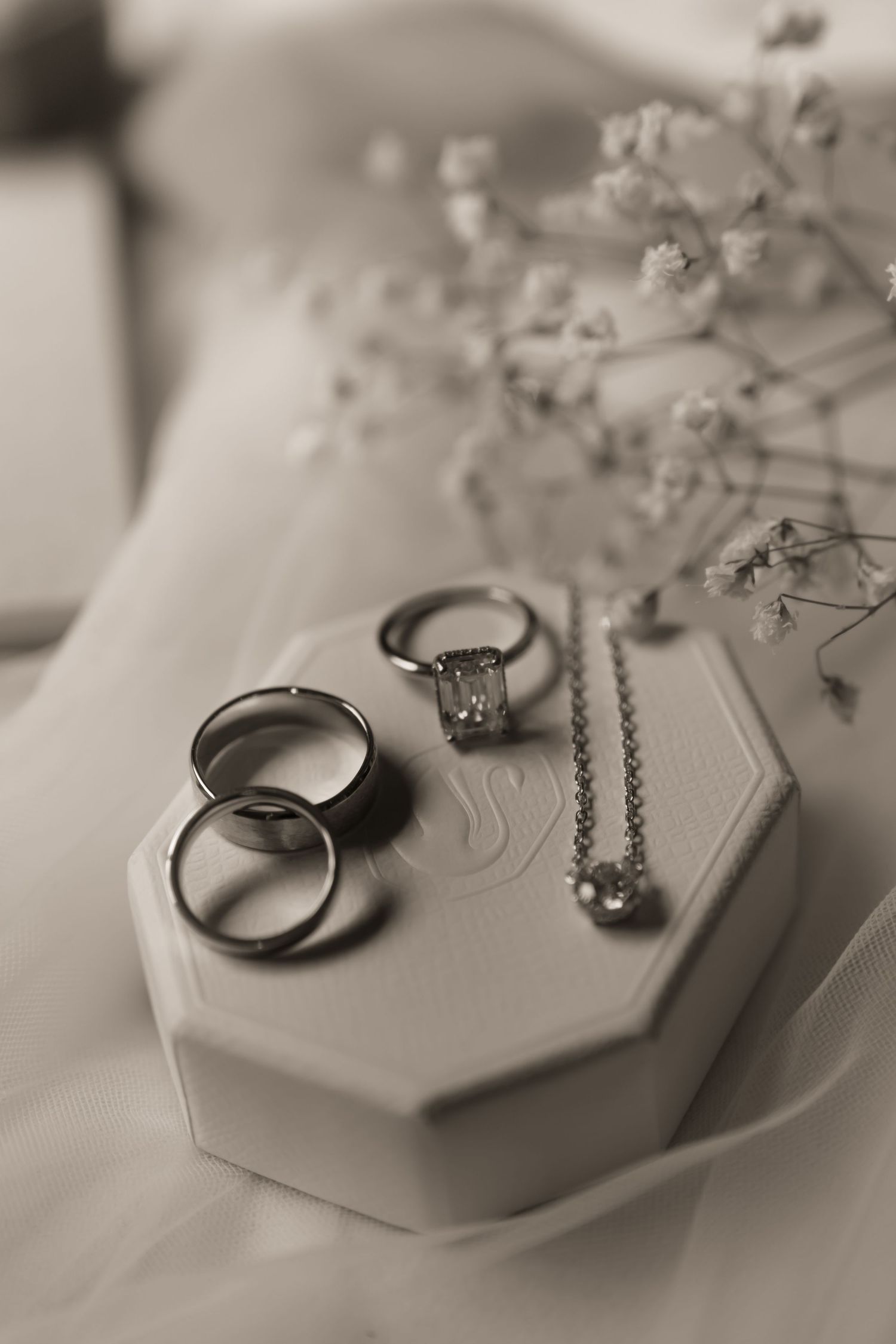 Black and white photo of wedding rings, diamond engagement ring, and necklace displayed on an octagonal box with baby's breath flowers.