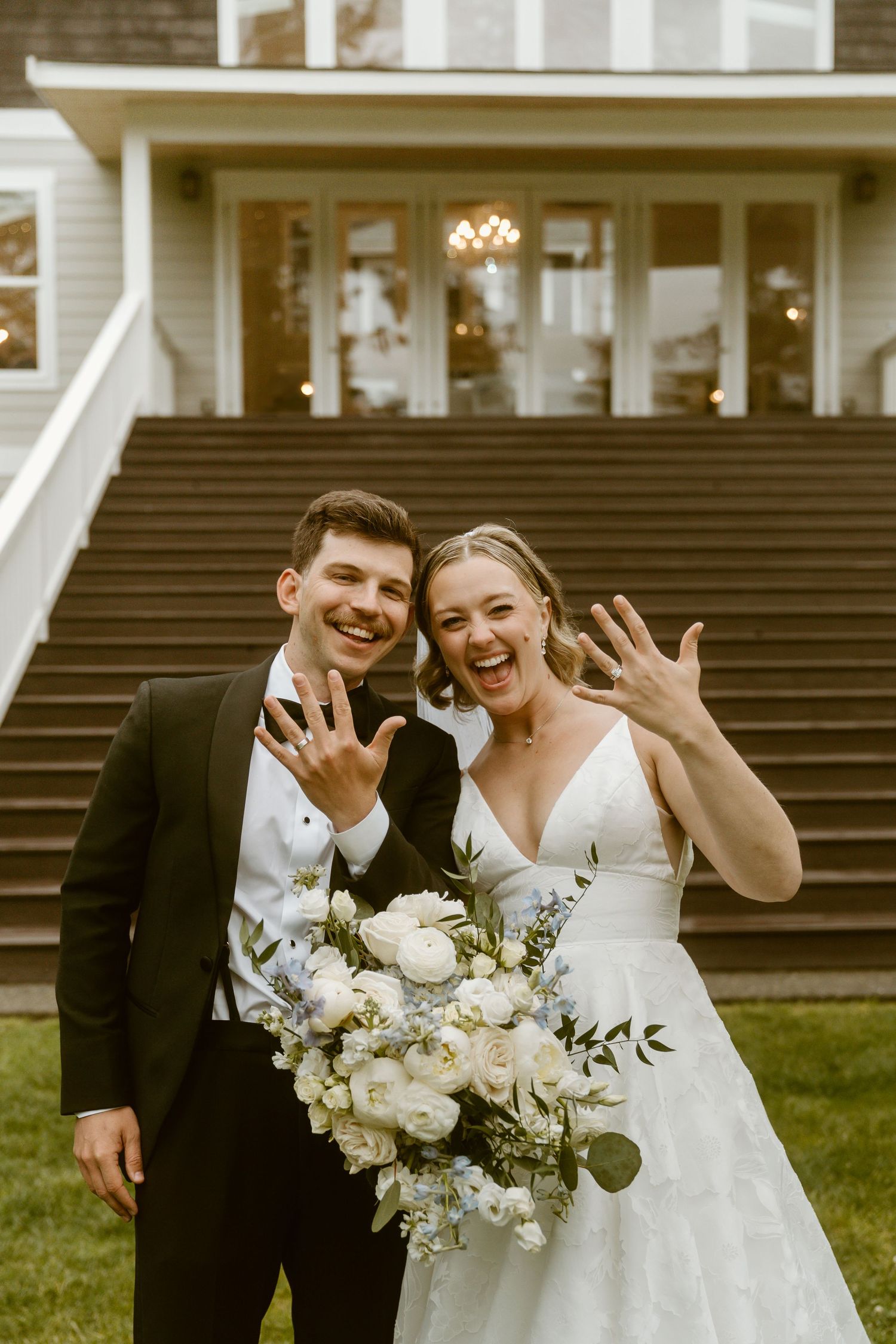 Joyful bride and groom showing off wedding rings outside elegant venue, bride holding white floral bouquet.