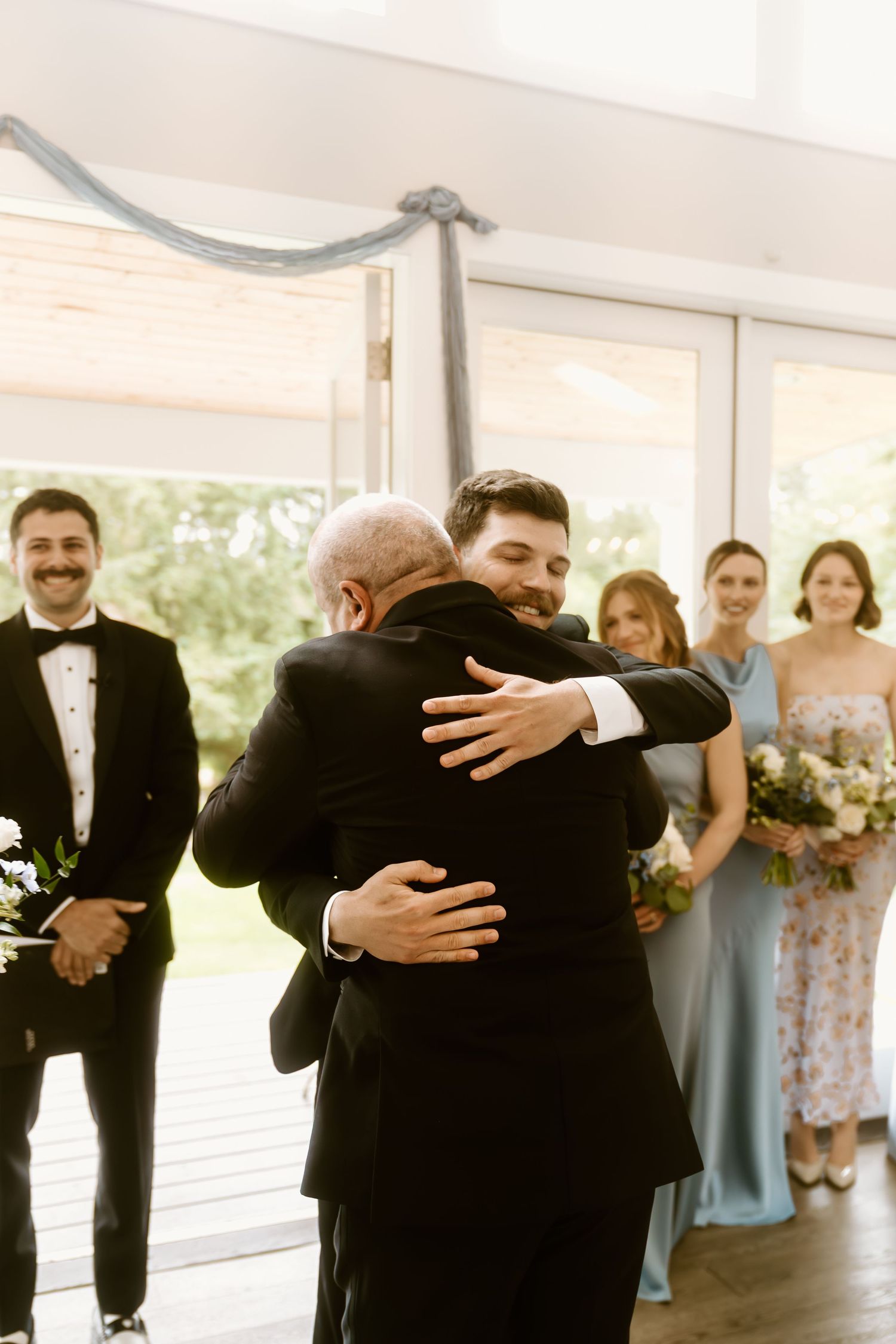 Groom shares emotional hug with father during wedding ceremony while bridal party looks on in elegant indoor venue.