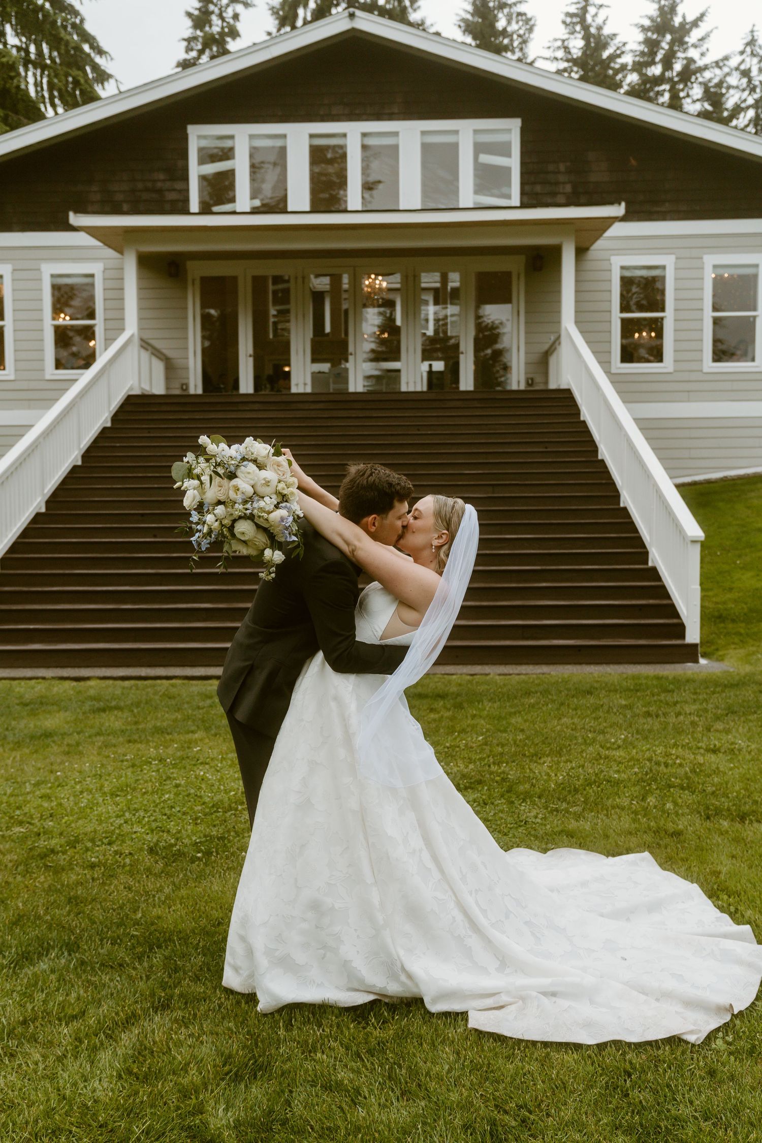 Groom dips bride for romantic kiss on green lawn in front of charming wedding venue.