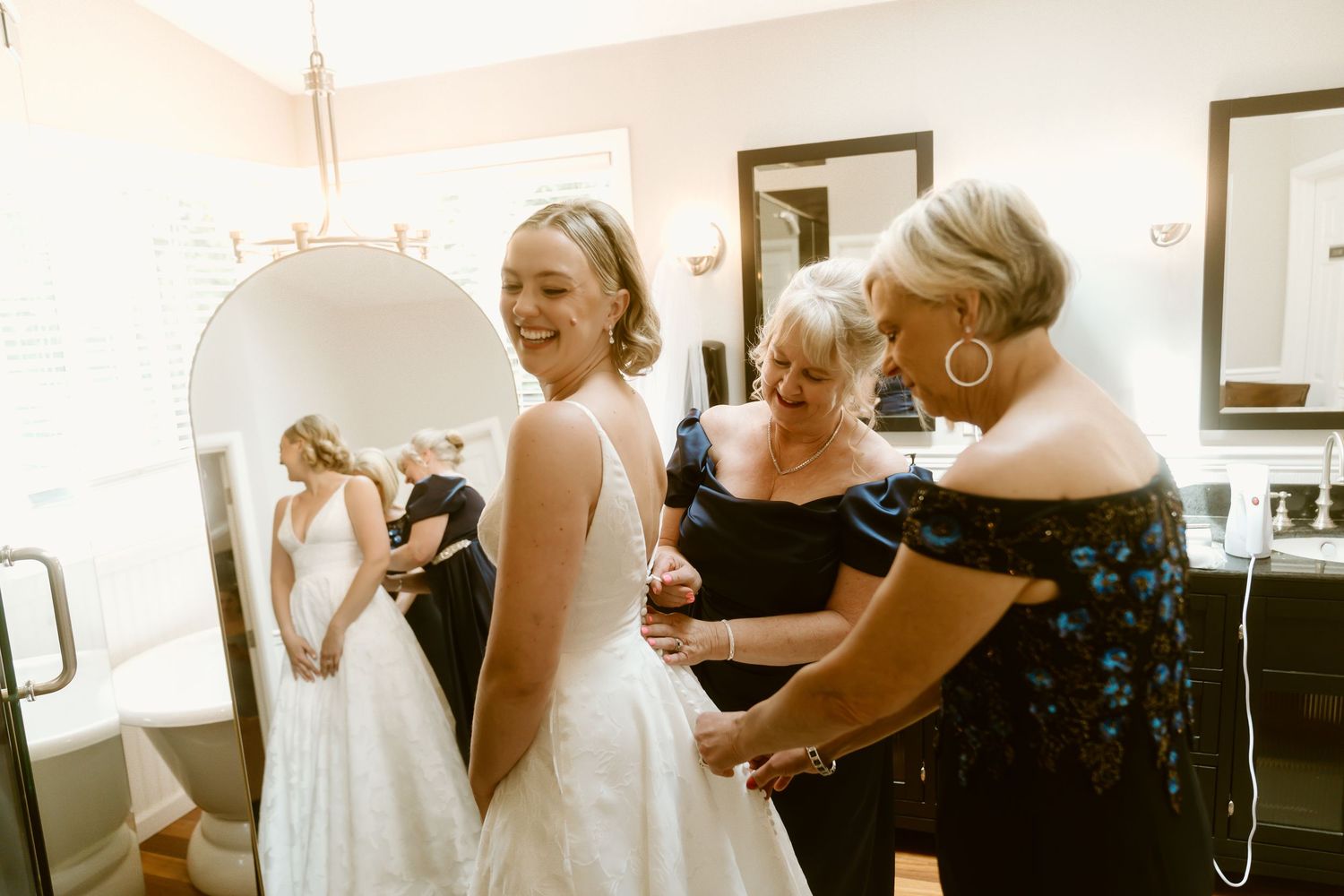Bride laughing as two women help fasten her white wedding gown in a bright dressing room.