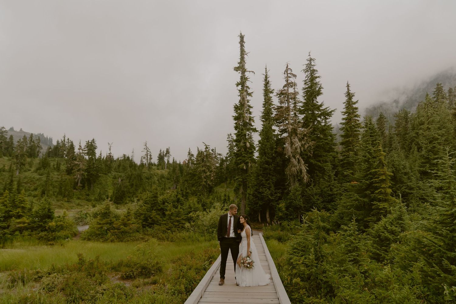 Couple walks on a wooden boardwalk surrounded by evergreen trees and misty mountains on their elopement day.