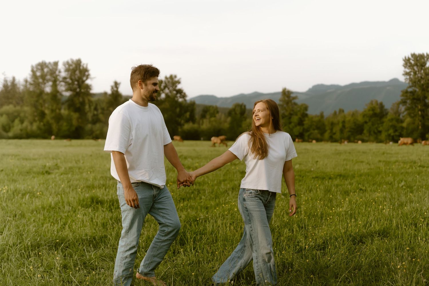 Couple holding hands walking in green field wearing white t-shirts and jeans with mountains in background.