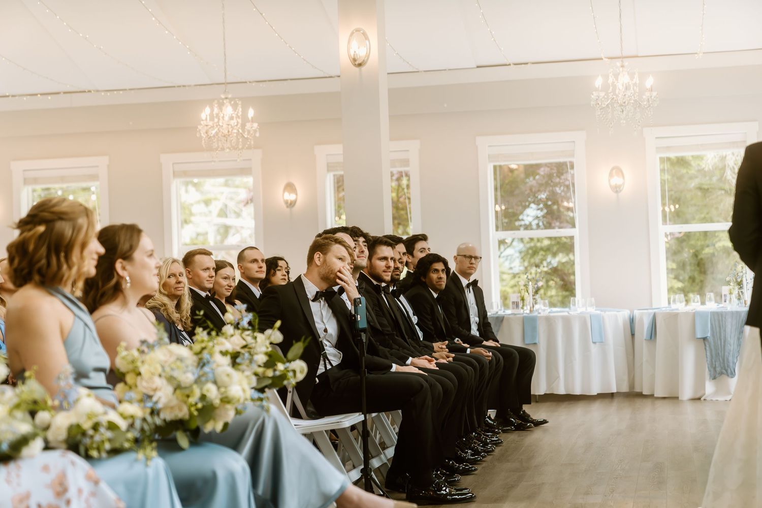 Guests in formal attire seated during an elegant indoor wedding ceremony with chandeliers and blue bridesmaid dresses.