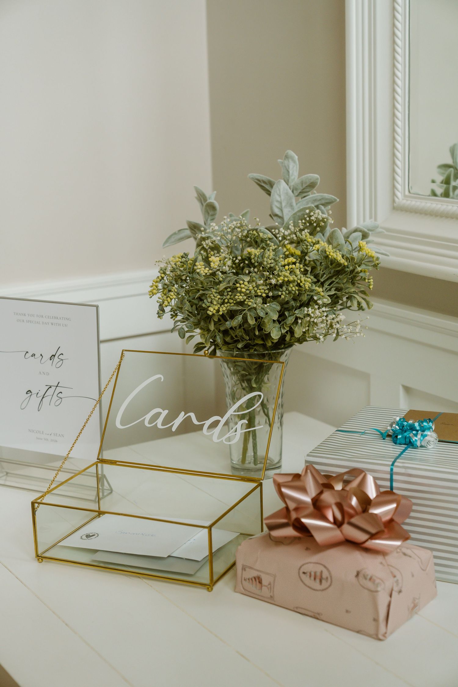 Gold glass card box with cards and gifts sign beside wrapped present with bow and floral arrangement on white table.
