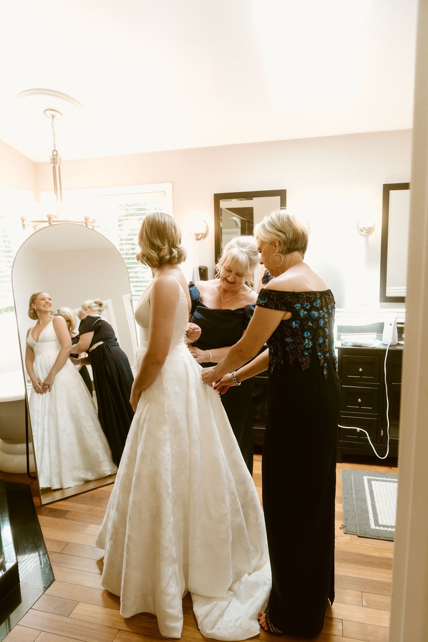 Bride in white gown being helped by two women in a elegant dressing room with a large arched mirror.