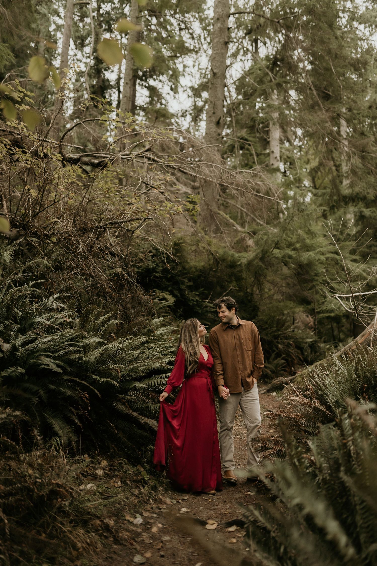 Couple holding hands on a forest path, woman in red dress, man in brown shirt, surrounded by ferns and tall trees.