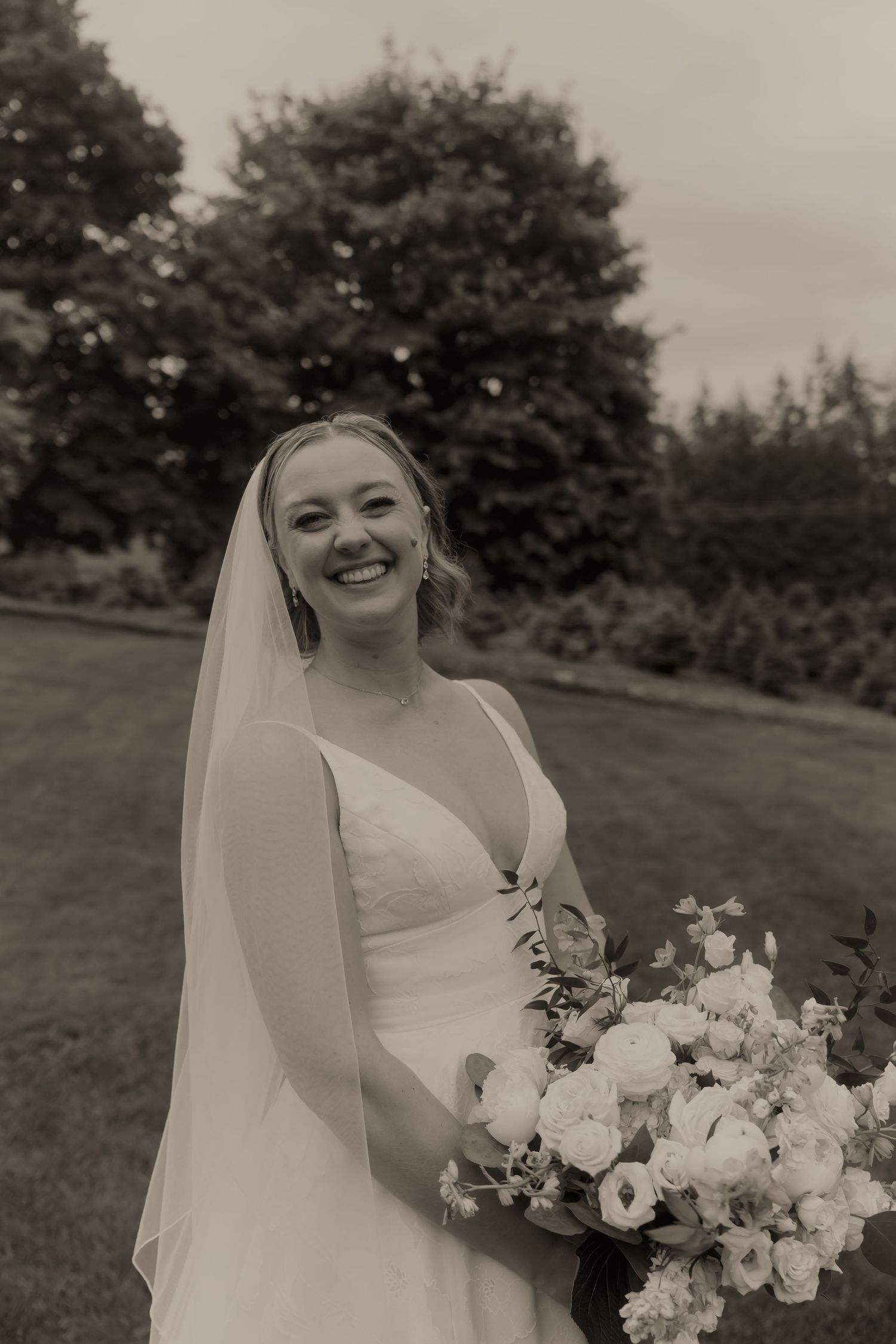 Smiling bride in white gown and veil holds bouquet of white flowers outdoors in black and white photo.