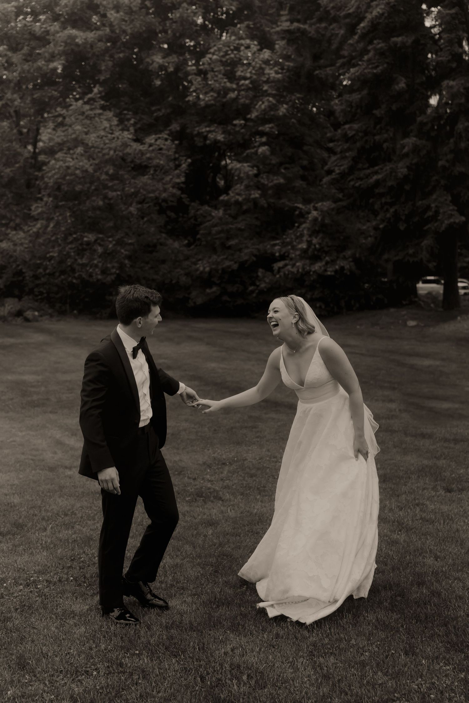 Black and white photo of a laughing bride in a flowing gown holding hands with a tuxedo-clad groom on a lush lawn.