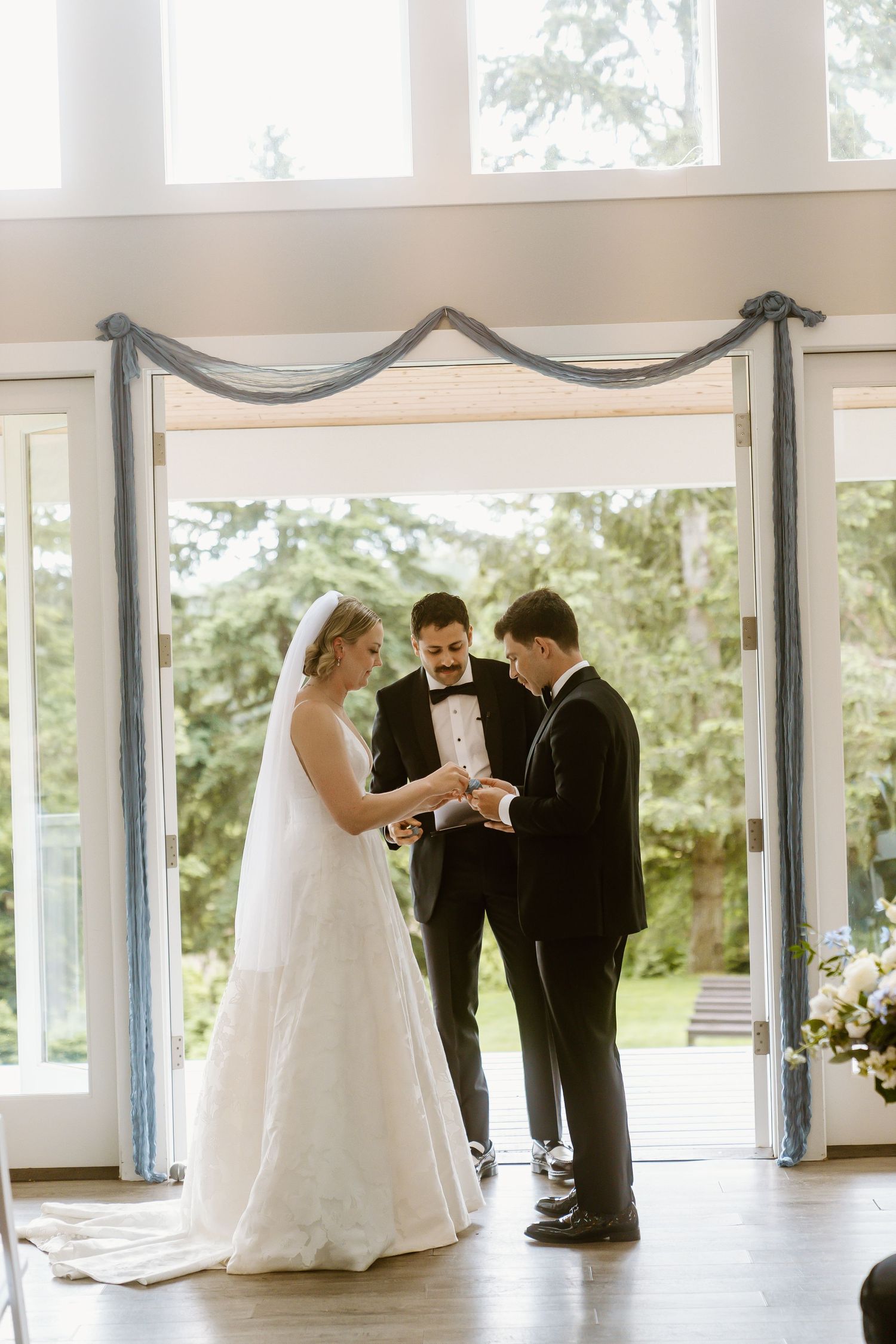 Bride and groom exchange rings during indoor wedding ceremony with officiant, framed by elegant French doors and draped blue fabric.