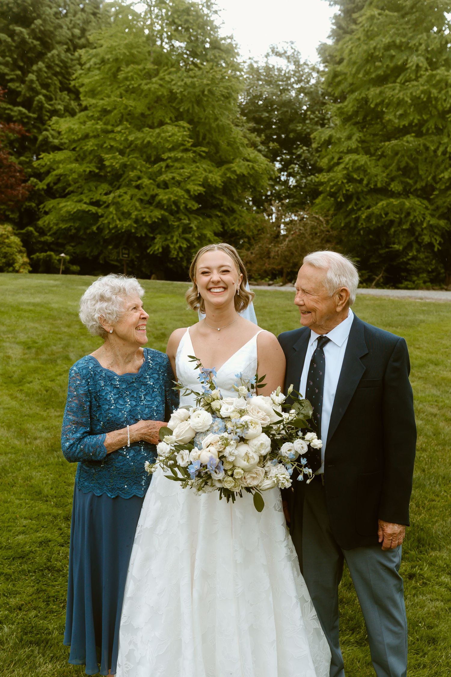 Smiling bride in white gown holding blue and white bouquet posing with elderly grandparents on wedding day.