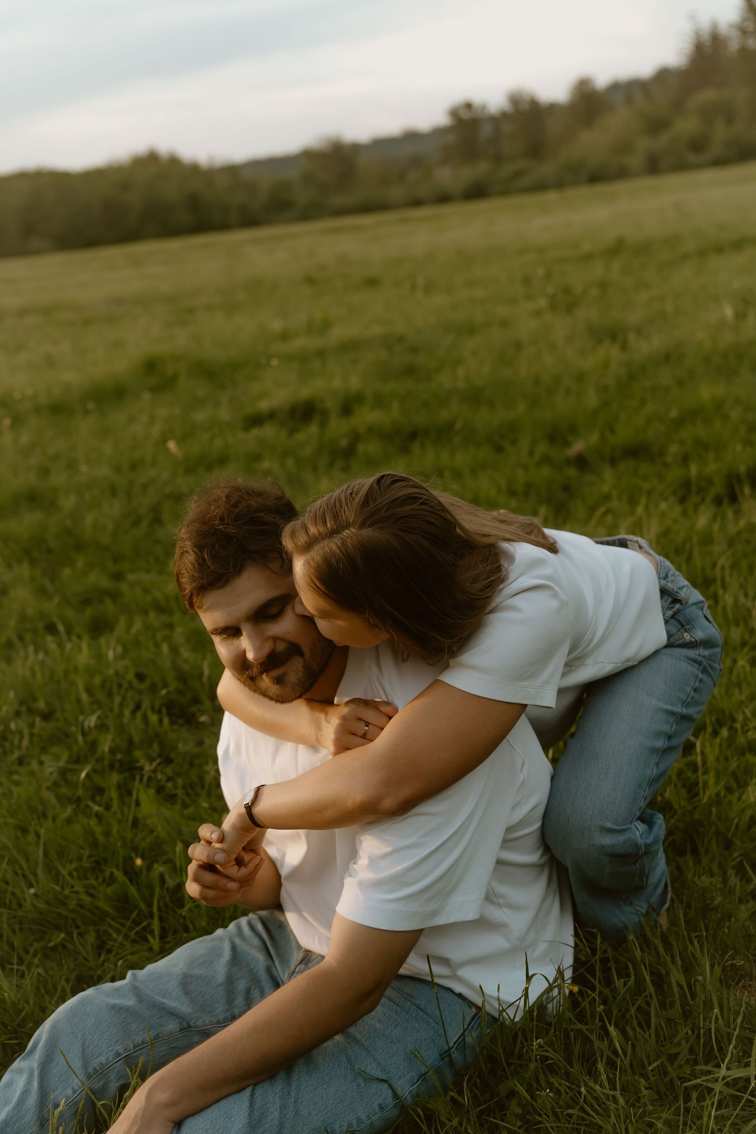 Romantic couple embracing on lush green meadow, woman hugging smiling man from behind at golden hour.