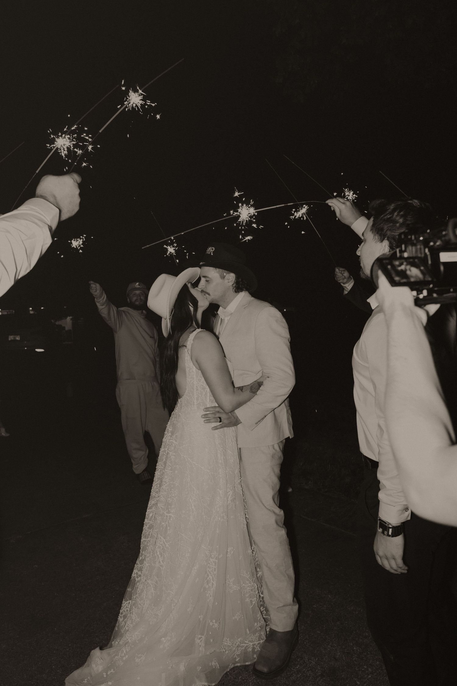 A couple shares a romantic moment at a nighttime wedding celebration while guests hold sparklers overhead.