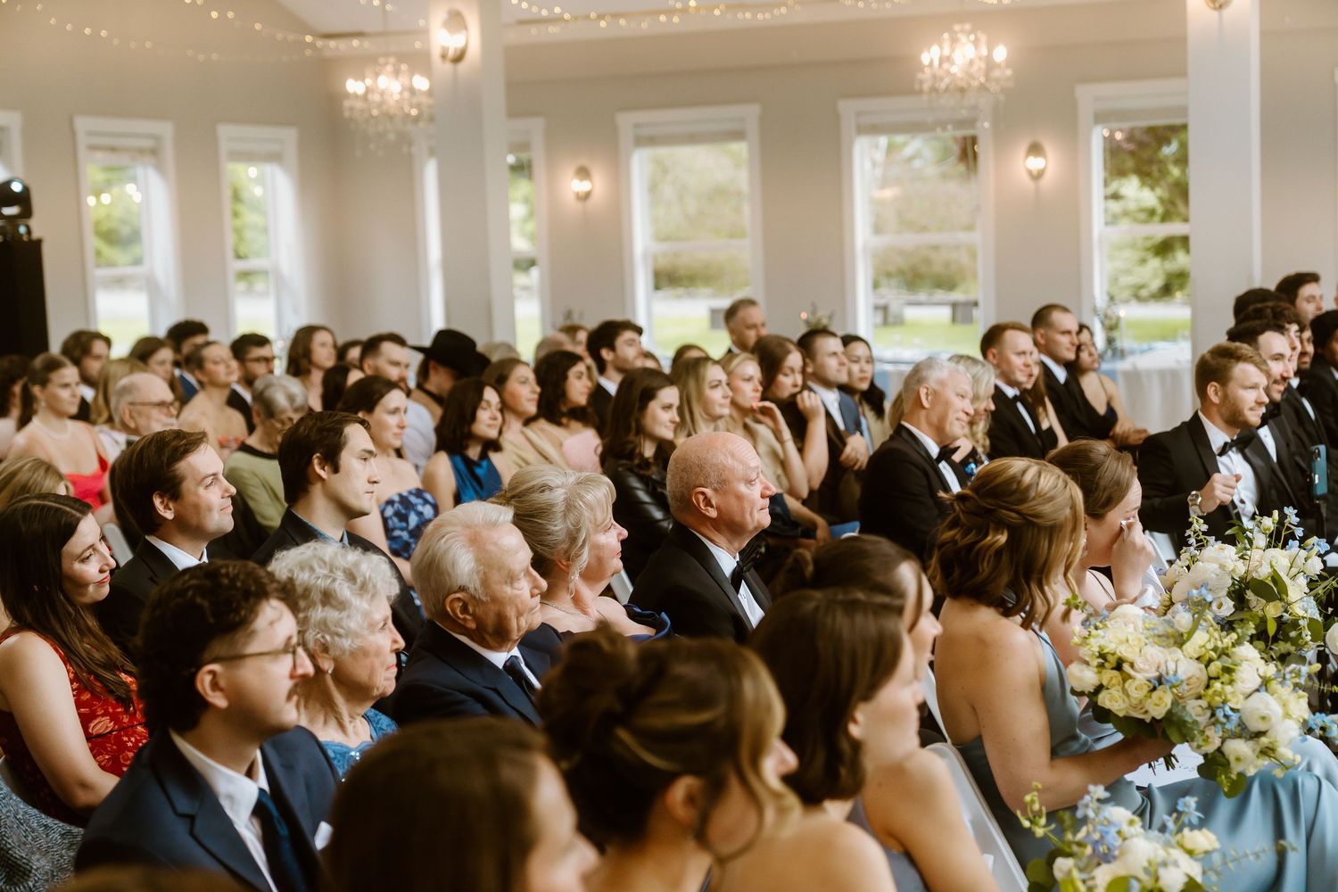 Elegant wedding reception guests seated in a bright ballroom, dressed in formal attire, smiling during the ceremony.