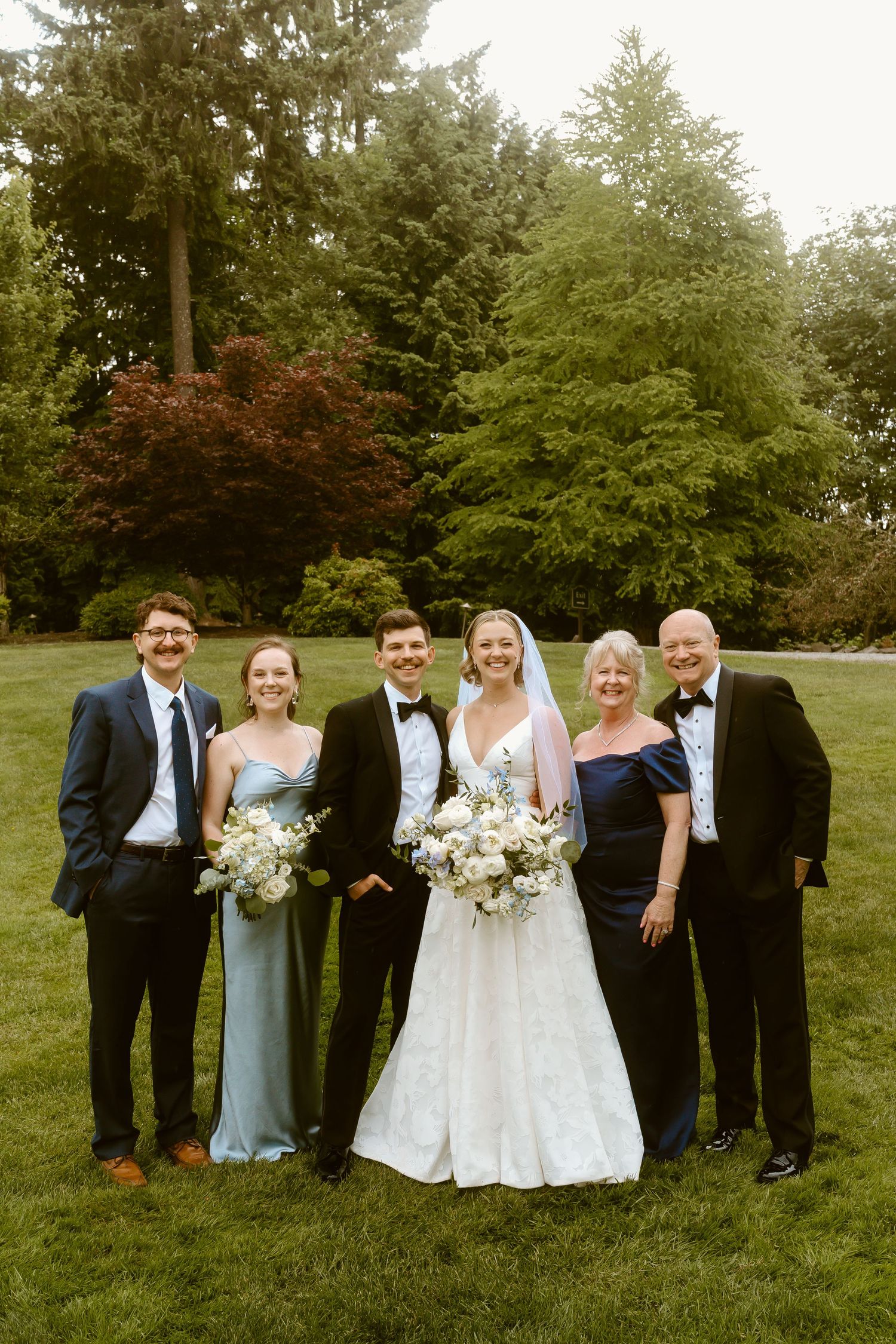 Wedding party family portrait with bride, groom, and four guests posing on a green lawn with trees behind.