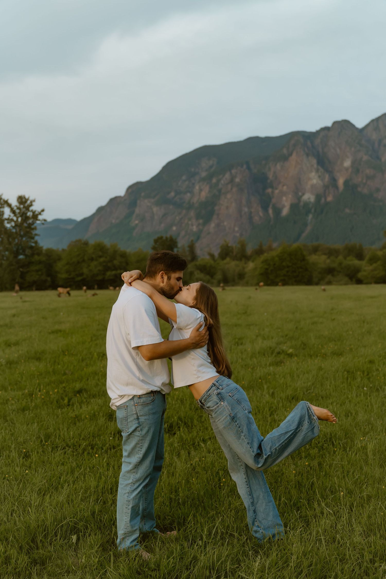 Couple sharing a romantic kiss in a green meadow with mountain backdrop, woman lifting her leg playfully.