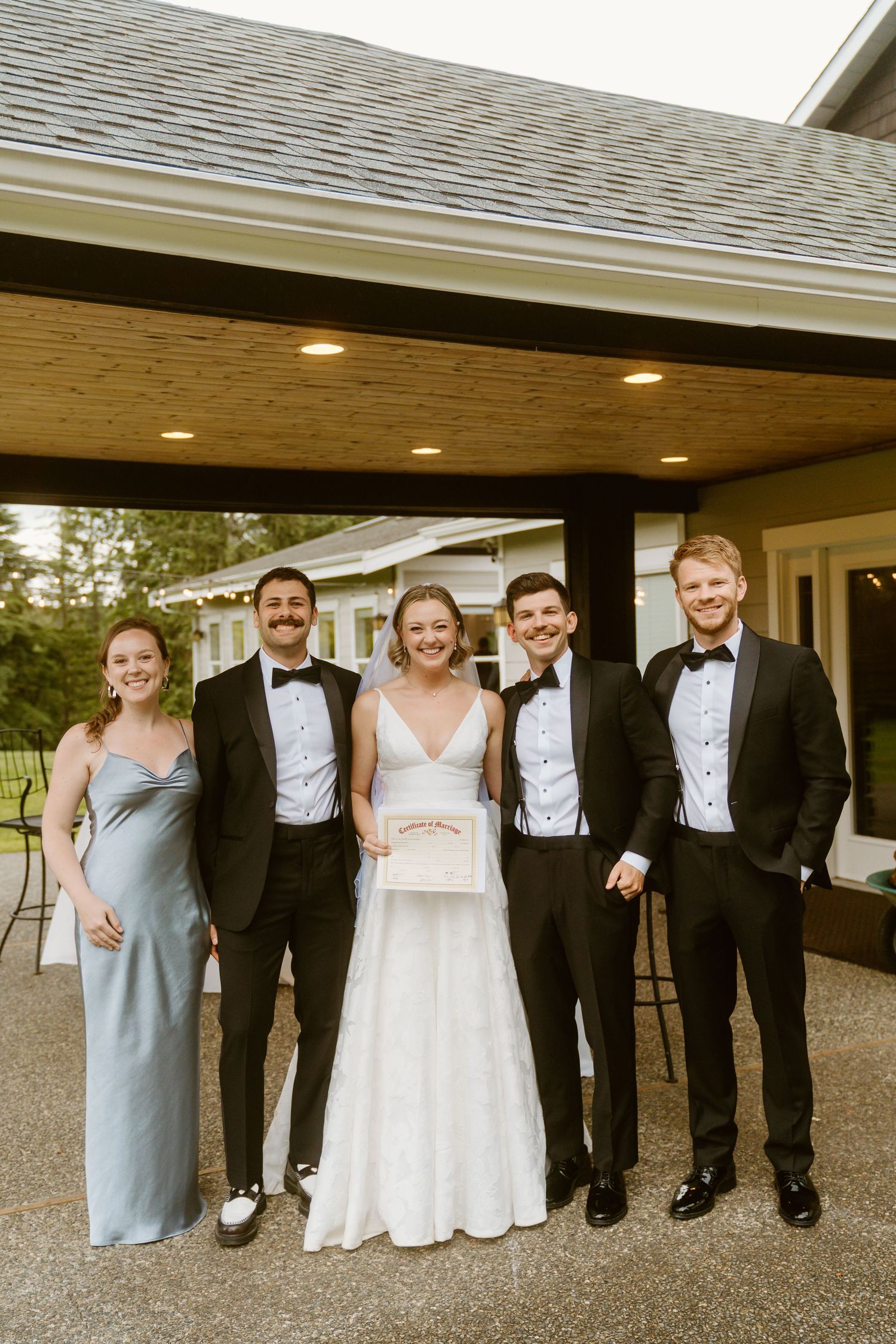 Bride holds marriage certificate while posing with wedding party outside venue after ceremony.