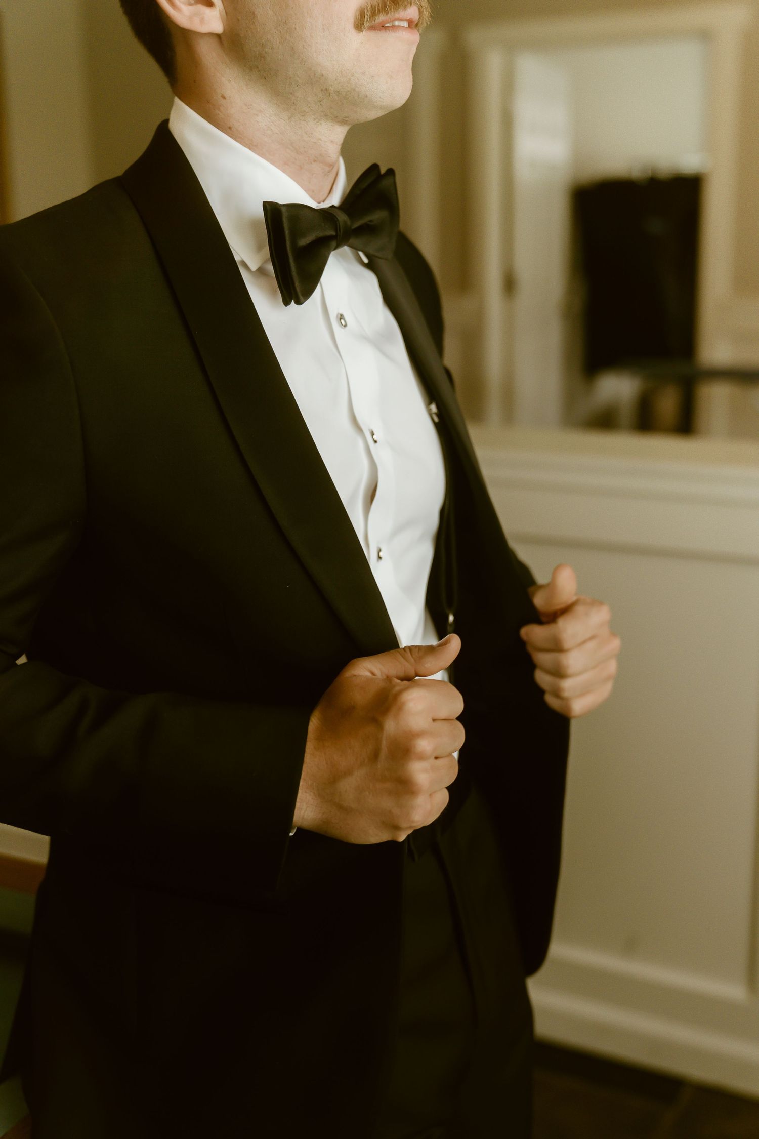 Groom in black tuxedo with bow tie adjusting his jacket lapels before wedding ceremony.