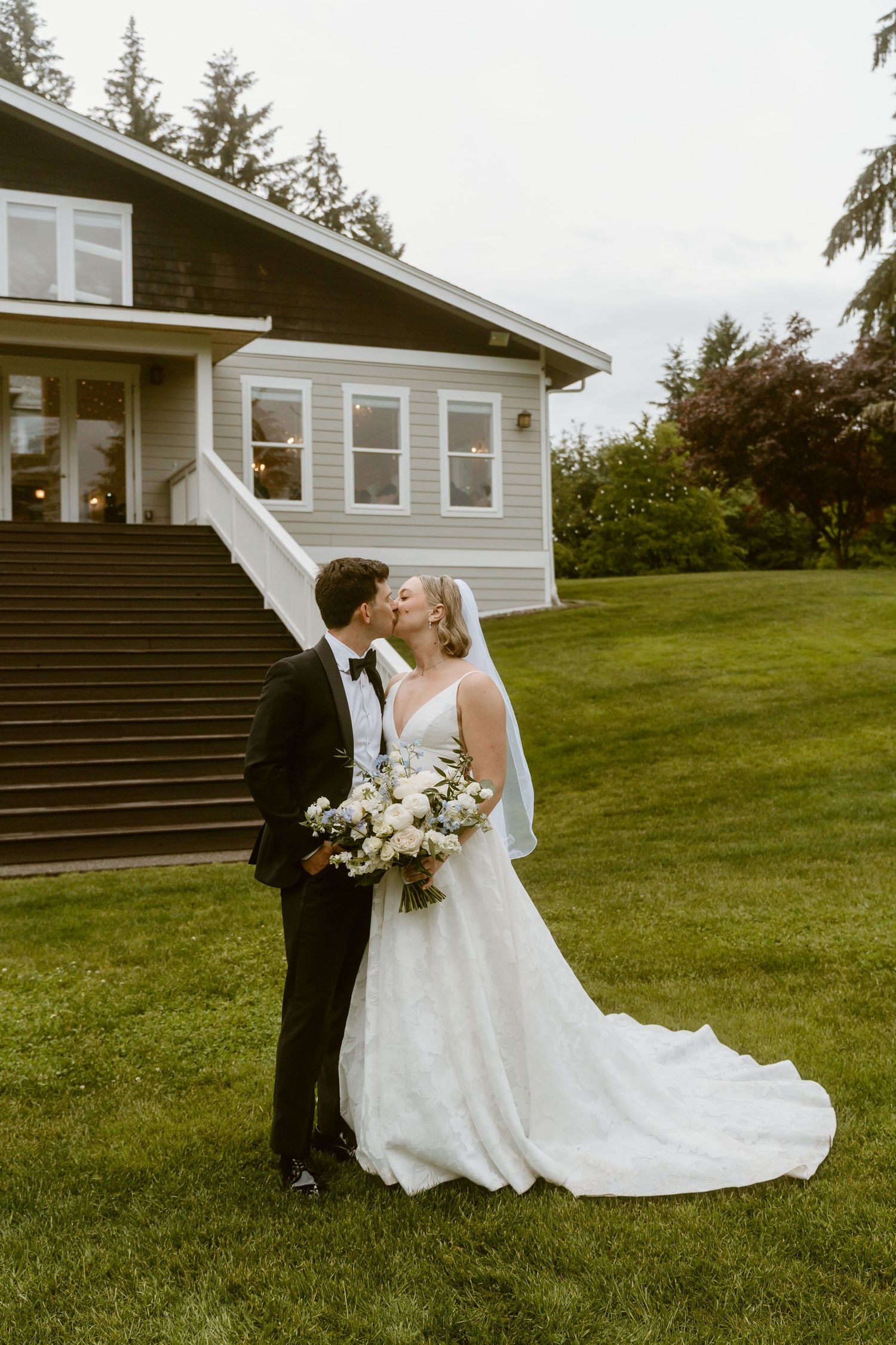 Bride and groom share a kiss on green lawn before a charming house, bride holding white floral bouquet in wedding gown.
