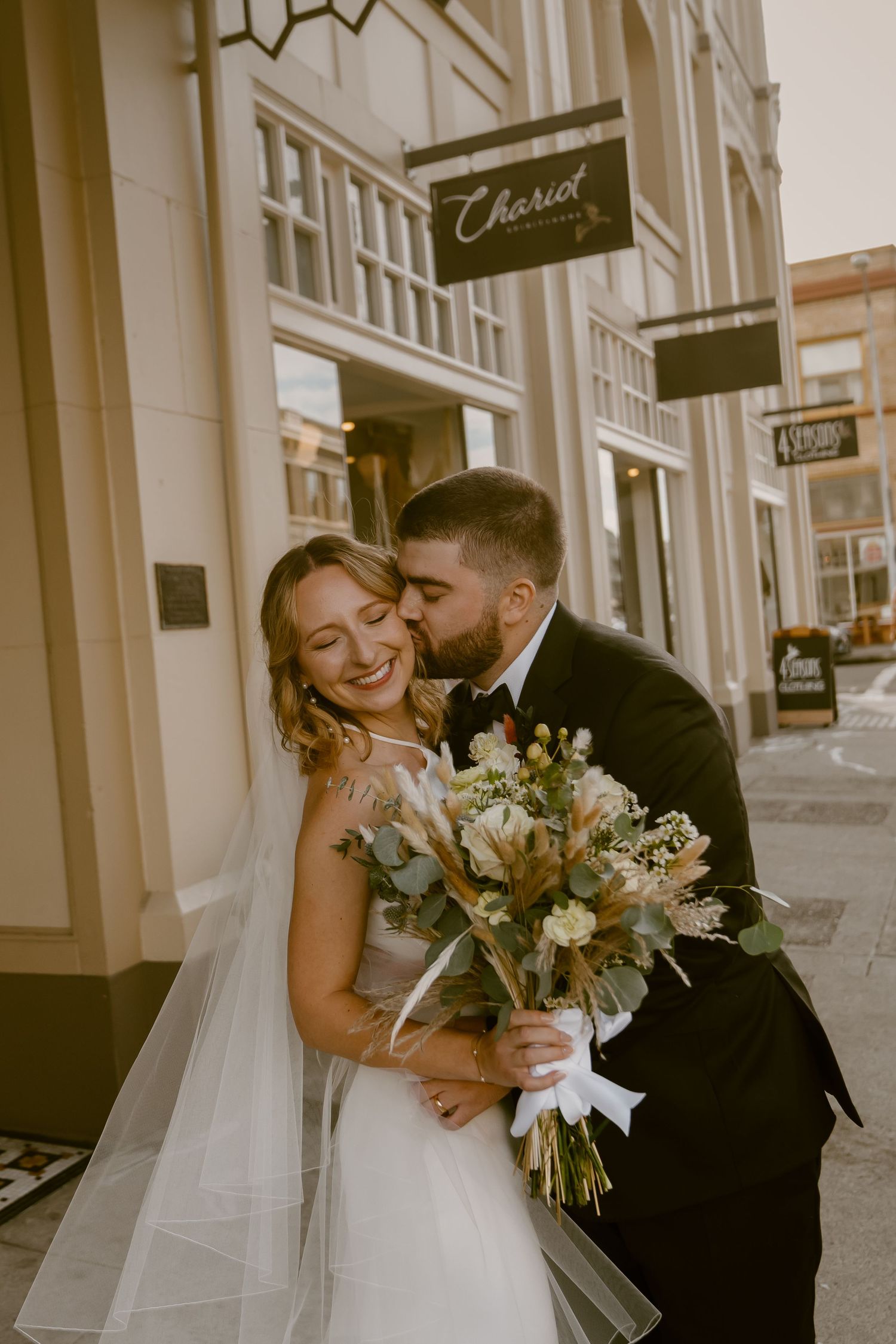 Newlywed couple shares romantic kisses on city street with bride holding elegant white and green floral bouquet.