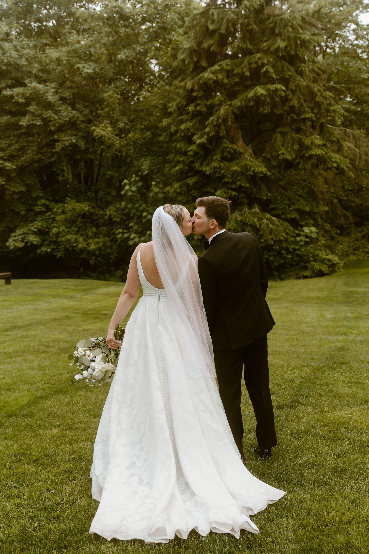 Bride and groom kissing in lush green garden, bride wearing flowing white gown with long veil.