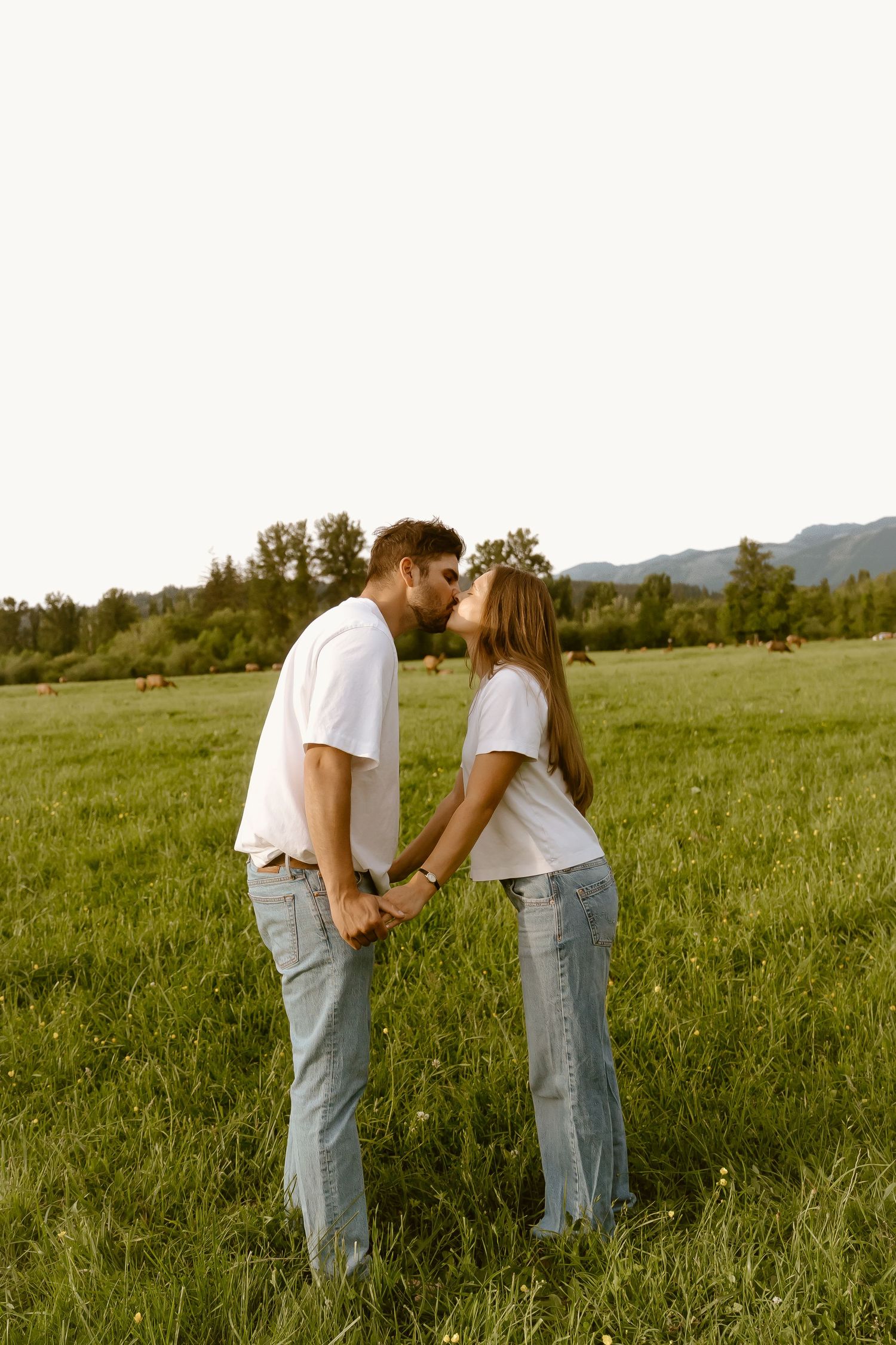 A couple in matching white tees and jeans share a kiss while holding hands in a lush green meadow.