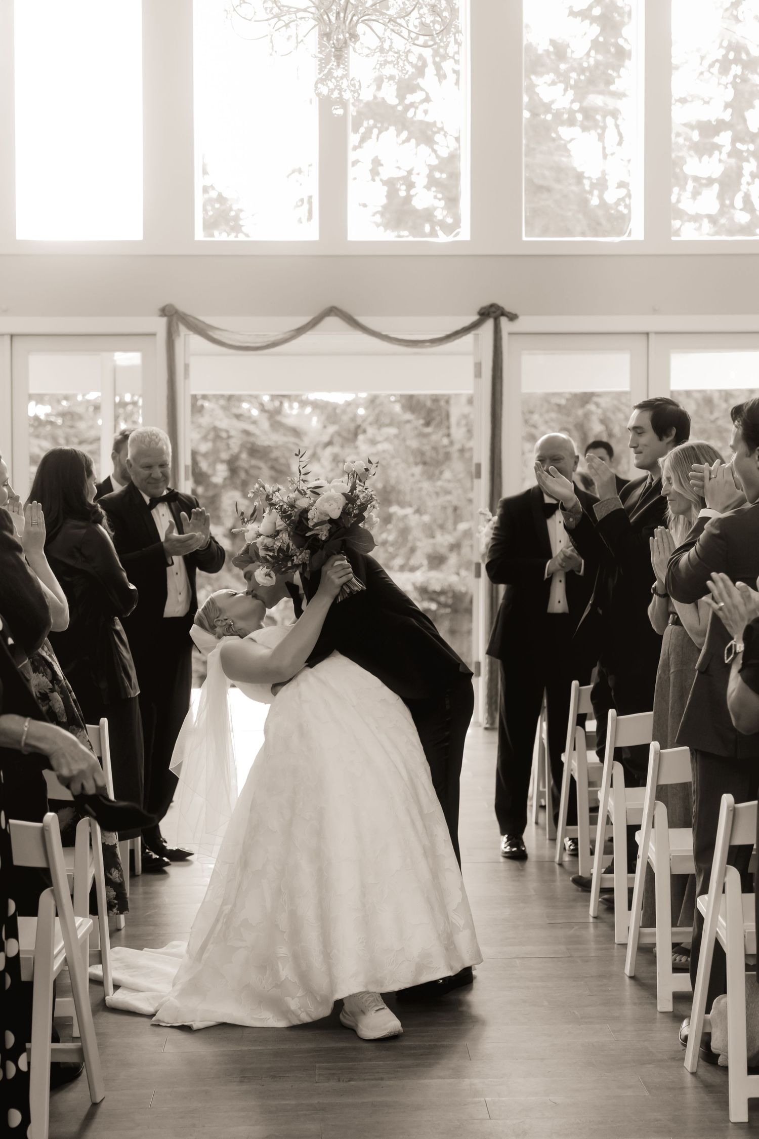 Black and white photo of newlyweds sharing a dip kiss while guests applaud in the aisle.