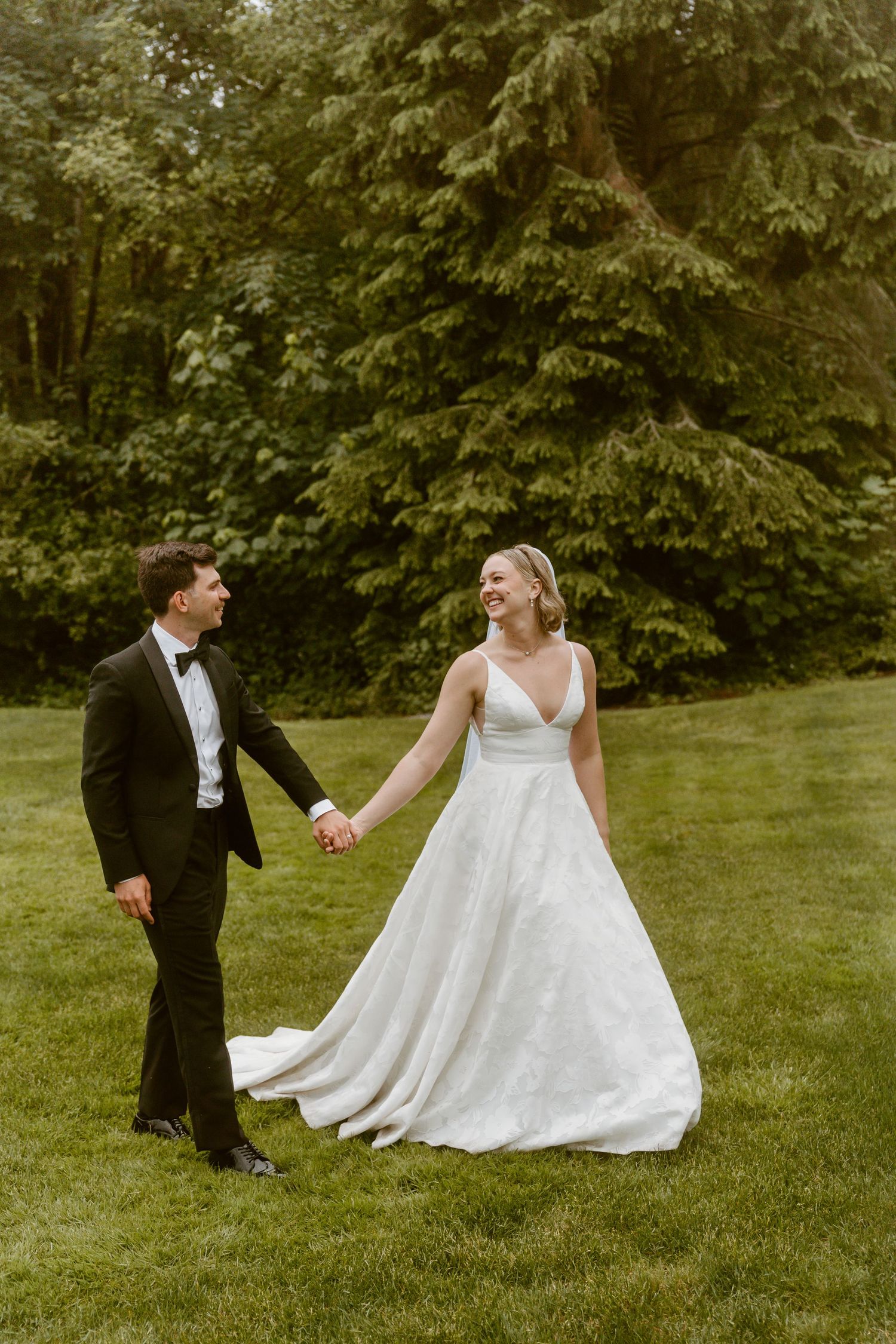 Bride and groom holding hands walking across lush green lawn with tall evergreen trees behind them.