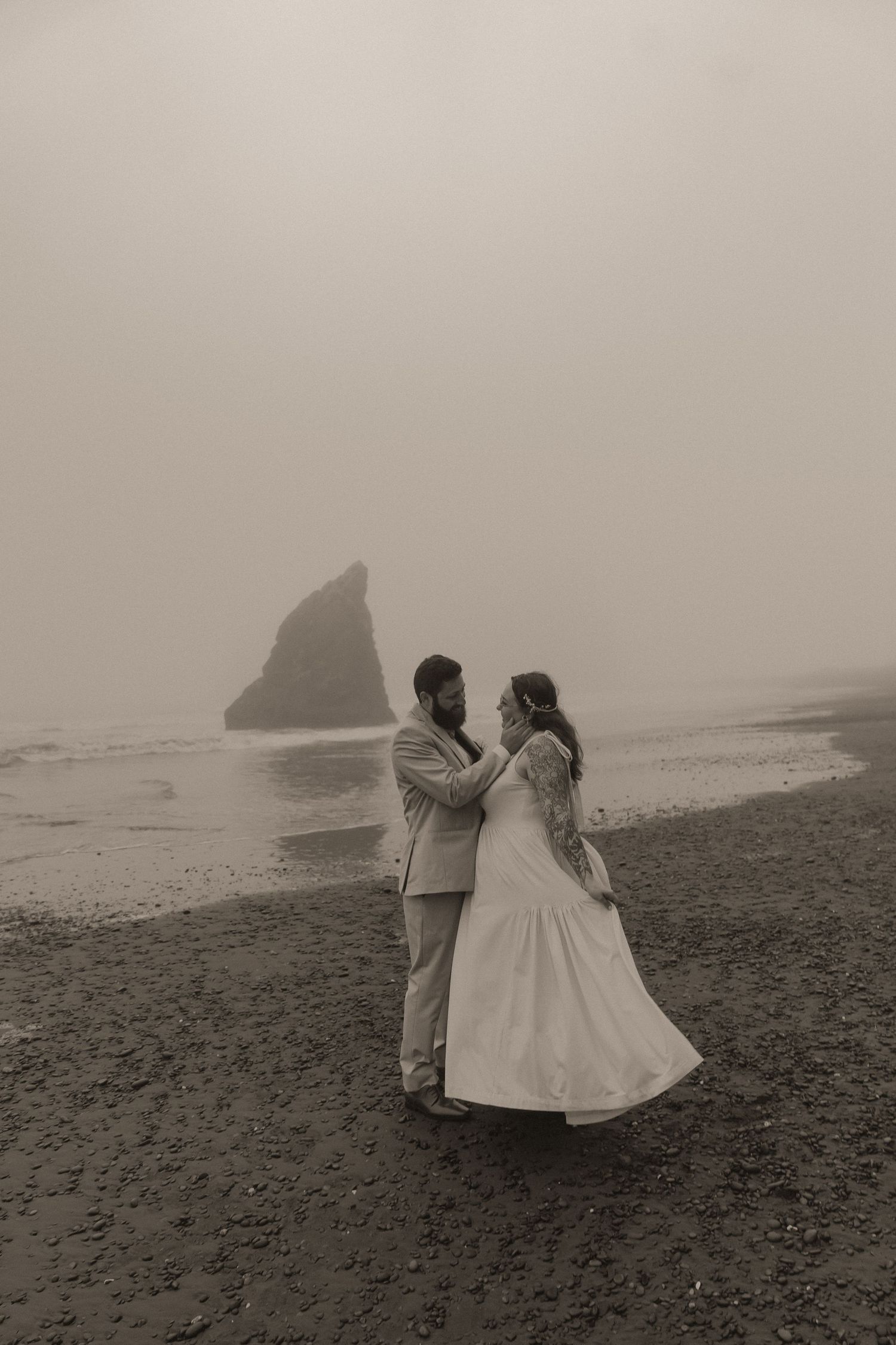 Couple in wedding attire embrace on a foggy, moody beach with a rocky sea stack in the background in a black and white elopement photo.