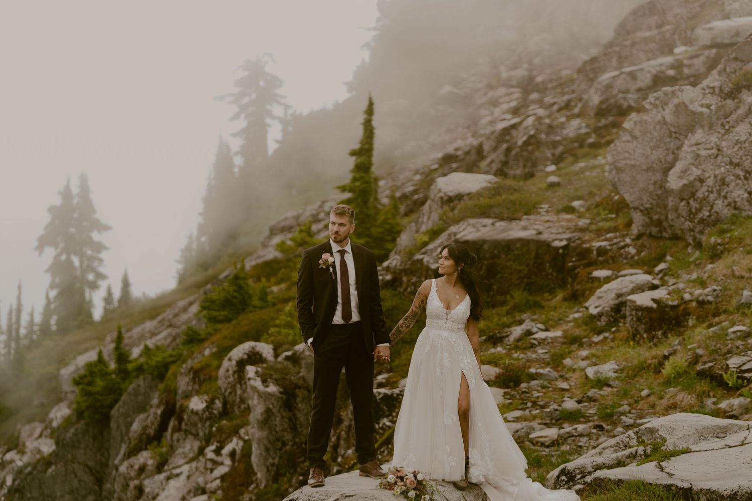 Bride and groom hold hands on a misty mountain rocky hillside surrounded by evergreen trees on their wedding day.