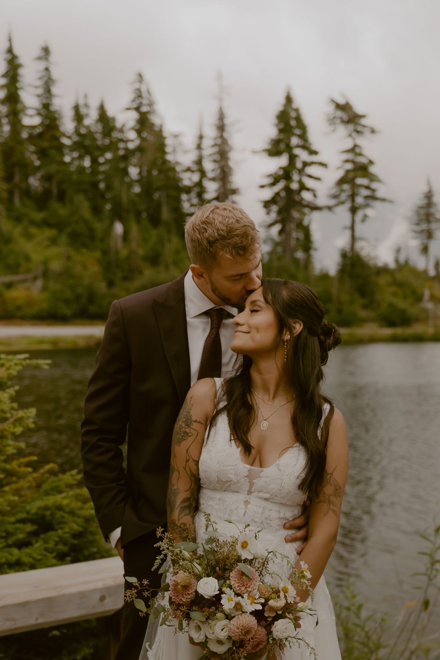 Romantic wedding couple embraces near a serene lake surrounded by evergreen trees in the Pacific Northwest.
