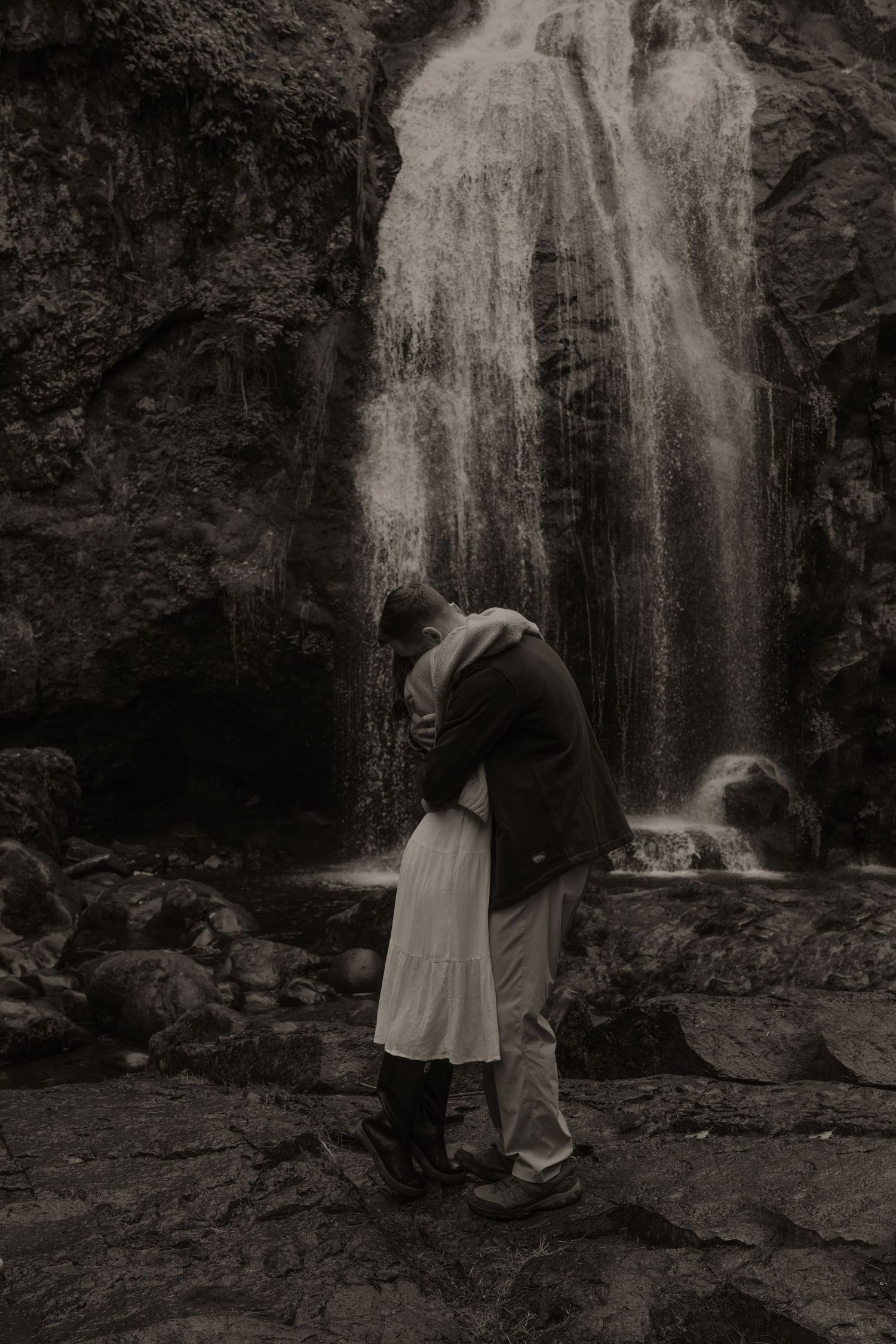 A romantic black and white photo of a couple embracing in front of a majestic waterfall.