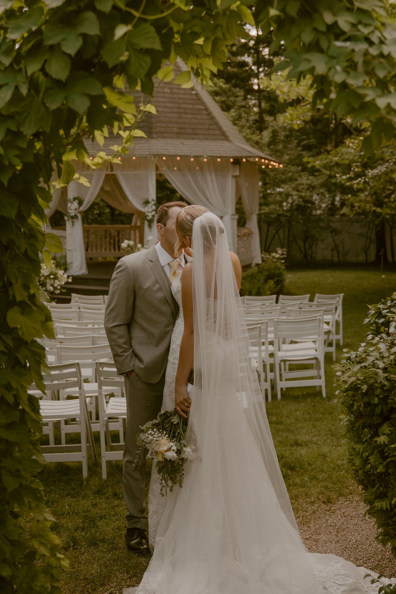 A romantic outdoor garden wedding ceremony setup with white chairs and a couple embracing under string lights and greenery.