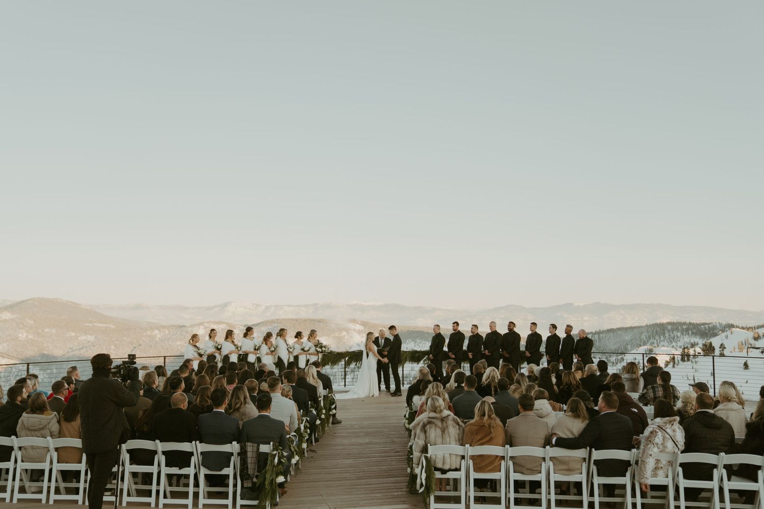 Outdoor waterfront wedding ceremony with white chairs, wedding party, and guests overlooking a scenic lake and mountains.