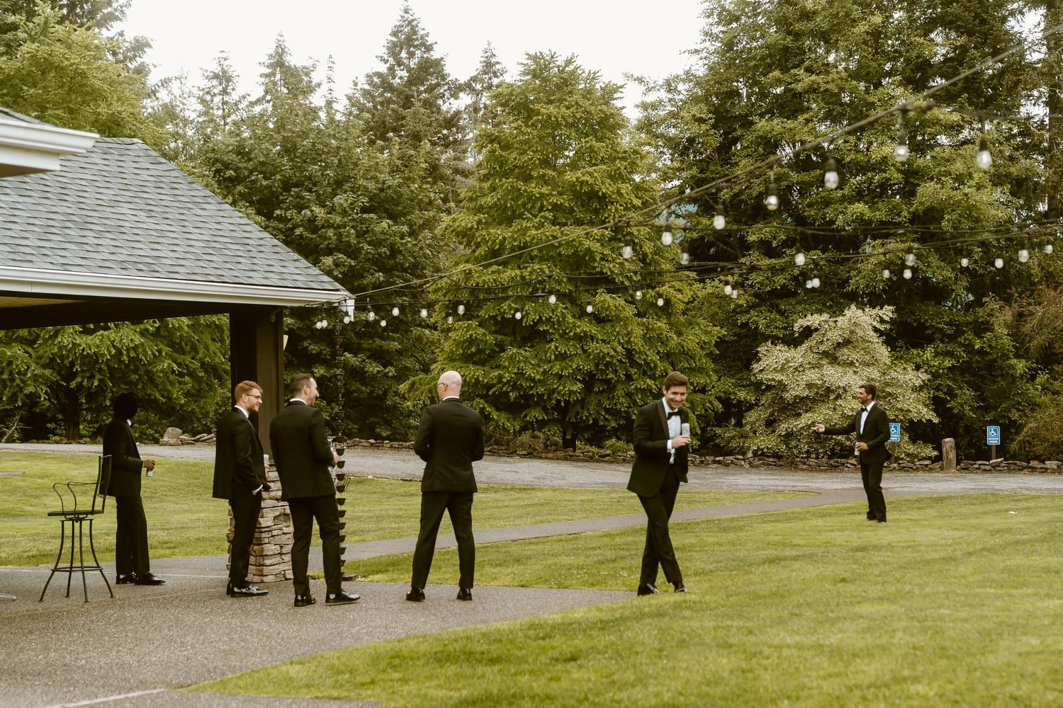 Groomsmen in black tuxedos gathered on a green lawn with string lights overhead.