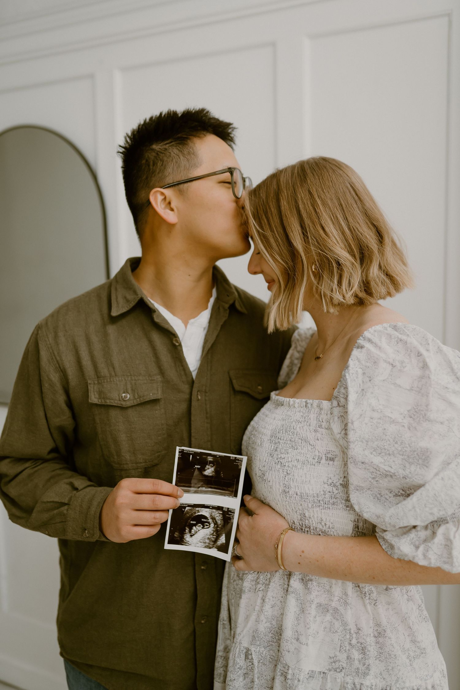 A couple in casual attire embraces while holding an ultrasound photo against a light gray background.