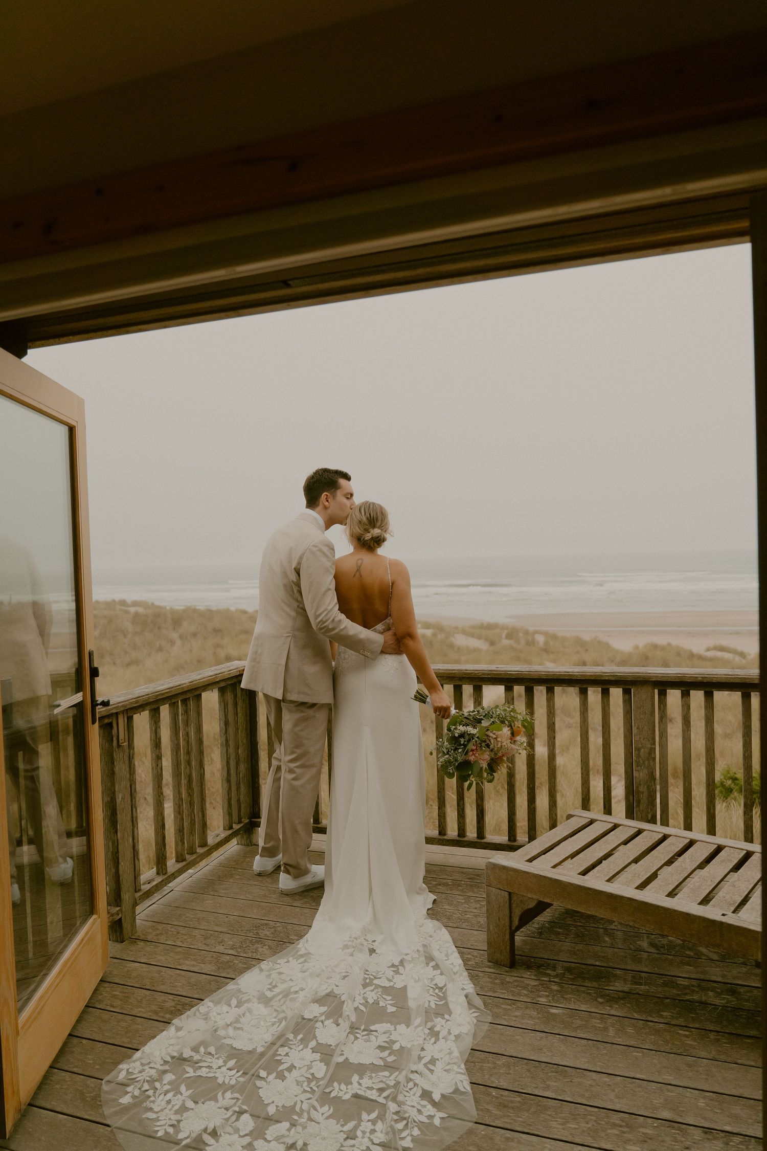 Wedding couple stands on wooden deck overlooking coastal beach landscape.