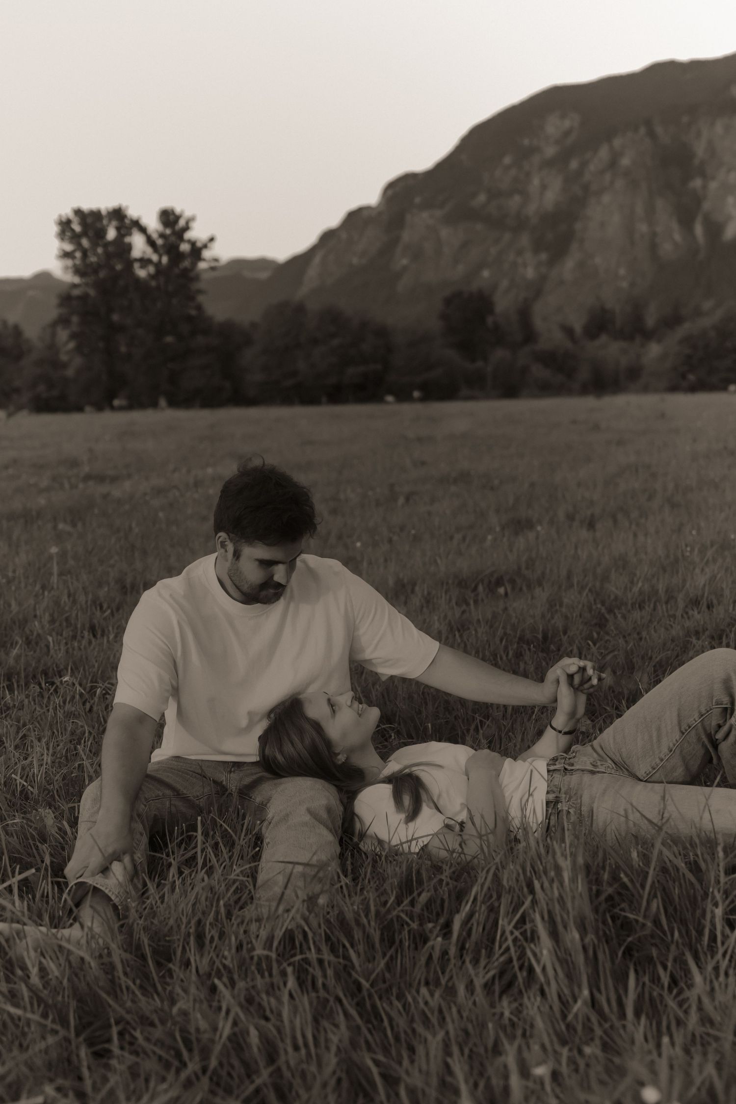 Couple sharing a romantic moment in a grassy field with mountains in the background, sepia-toned photo.