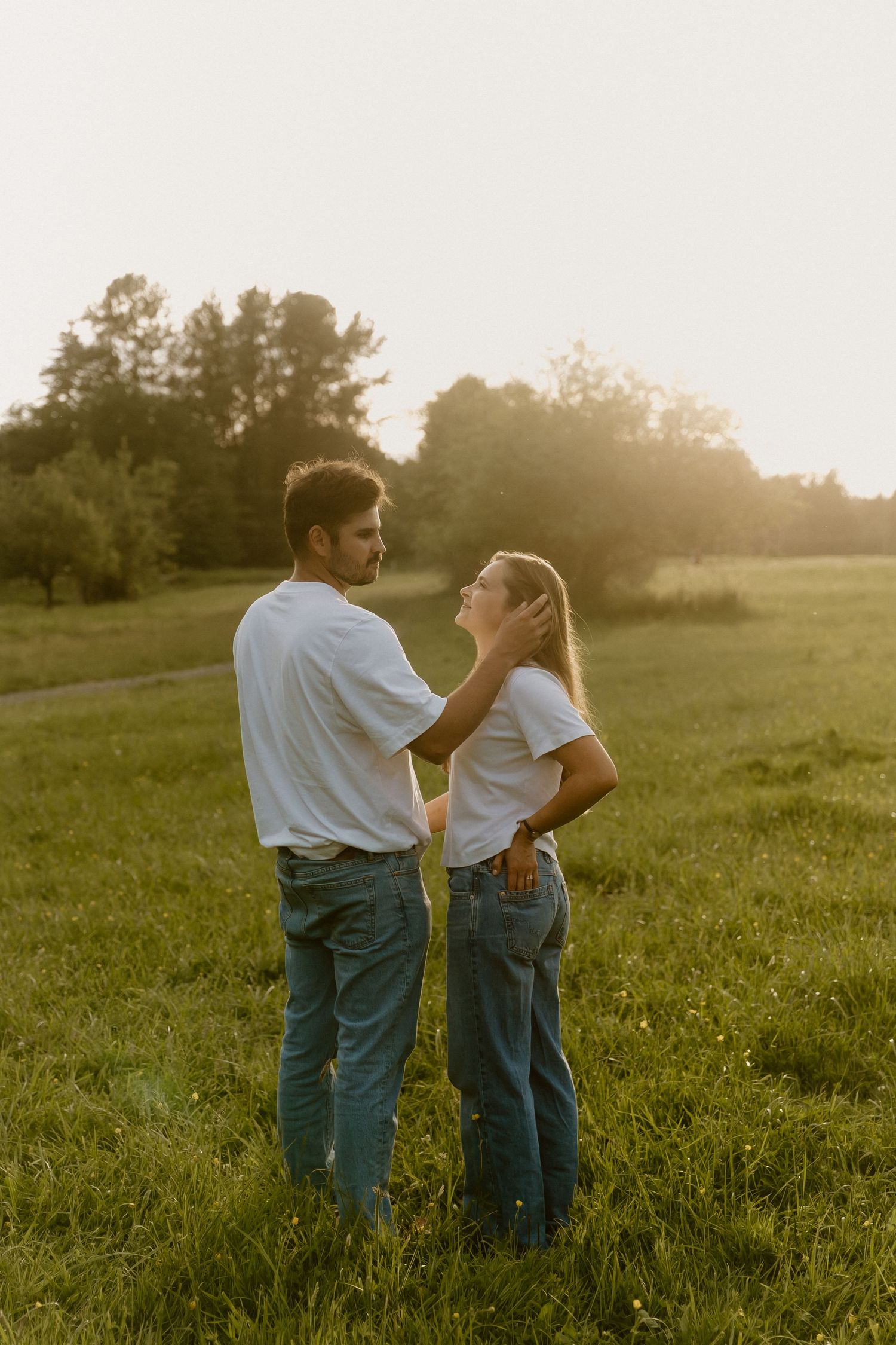 Couple standing in golden sunset light on green field, man gently touching womans face affectionately.