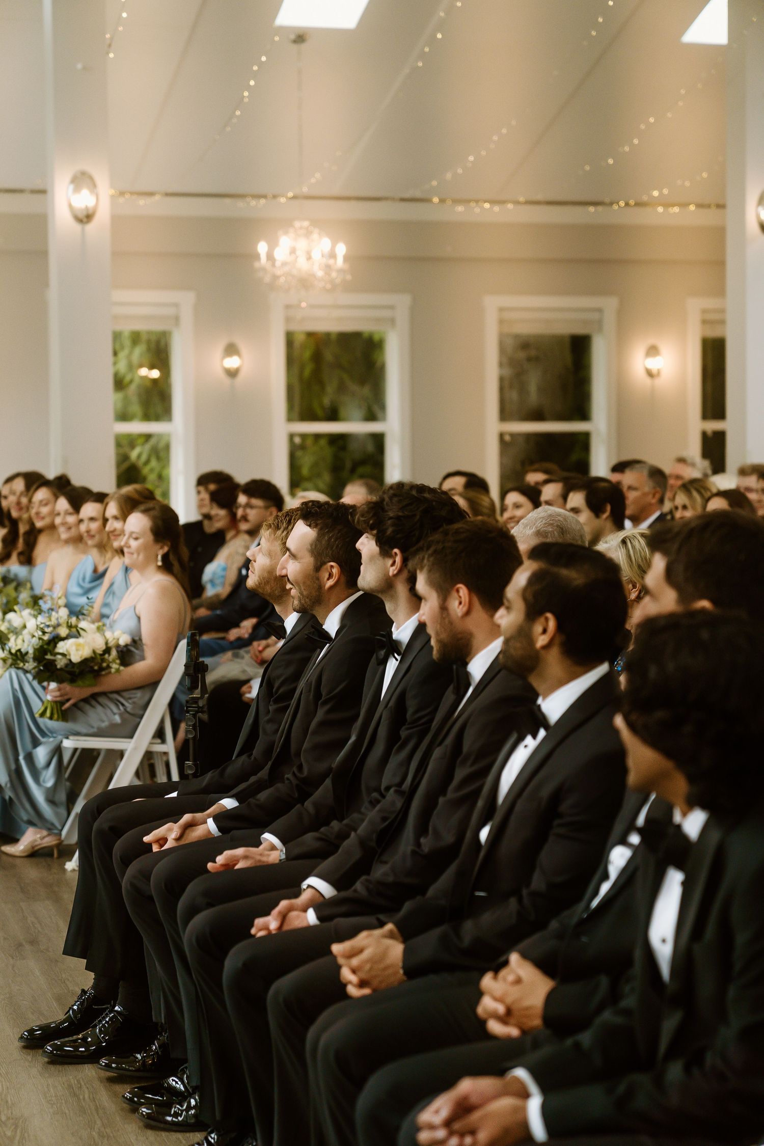 Groomsmen in black tuxedos seated in white chairs during an elegant indoor wedding ceremony.