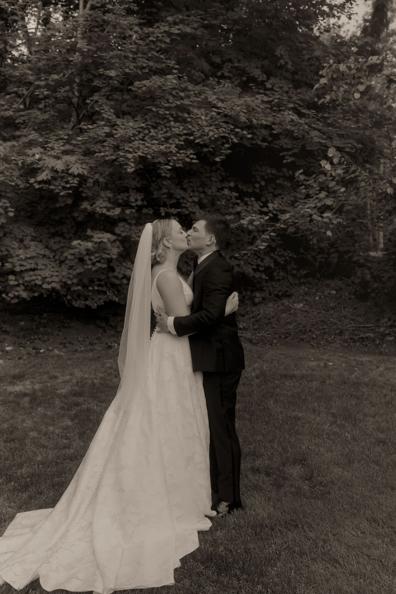 Black and white photo of bride and groom sharing a romantic kiss outdoors with lush trees in the background.