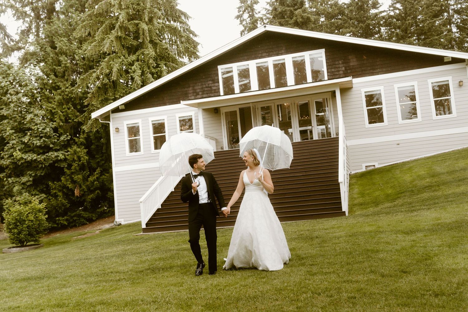 Bride and groom walking hand in hand on green lawn holding white parasols in front of elegant white wedding venue.