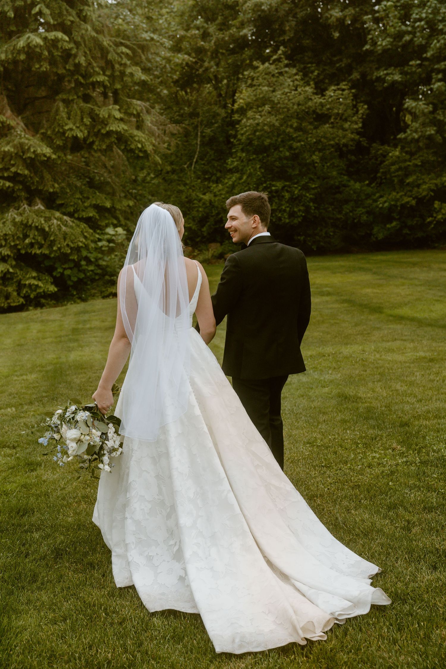 Bride and groom walking hand in hand through green garden, bride in elegant lace gown.