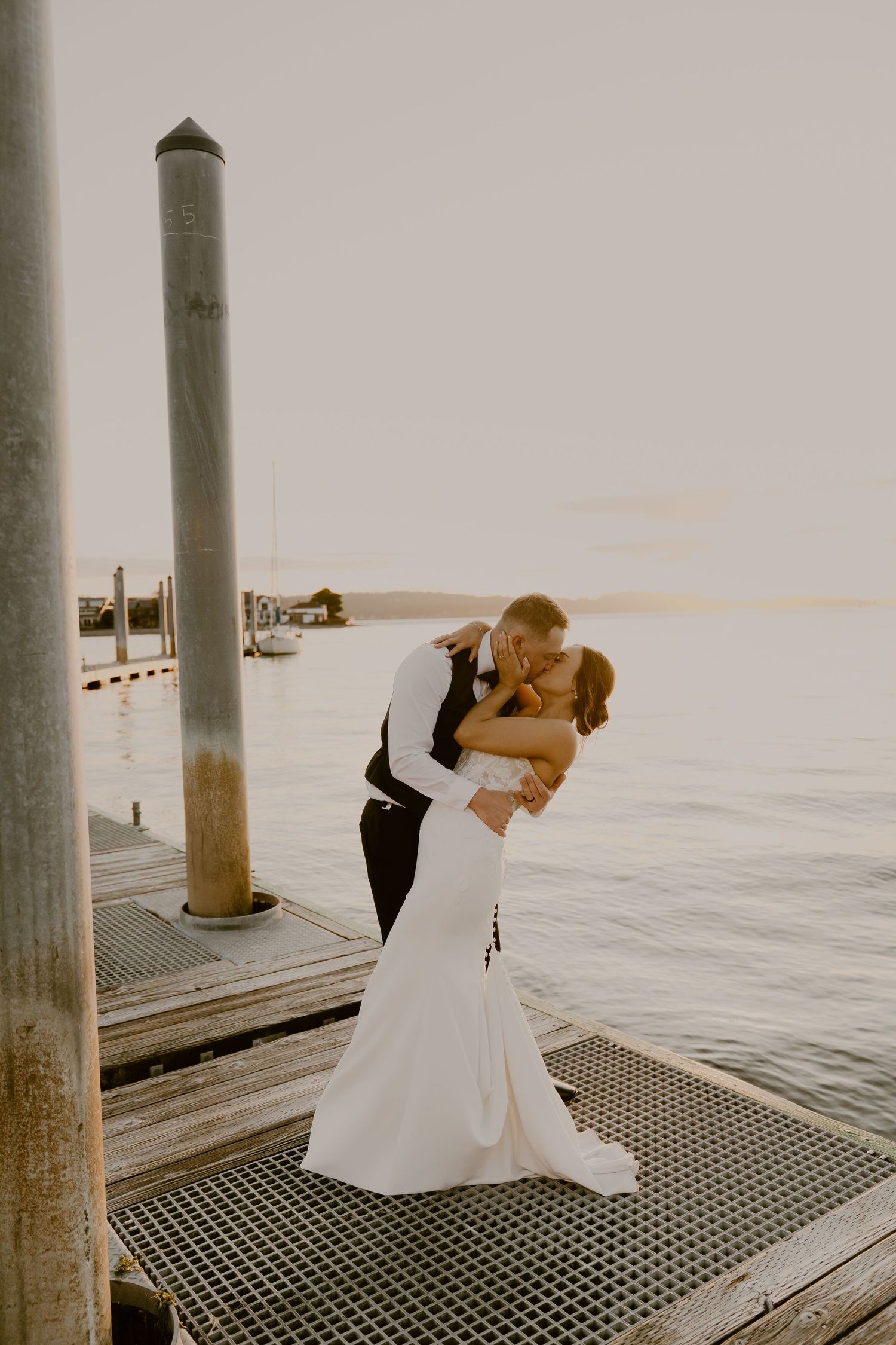 Bride and groom share a romantic kiss on a waterfront dock at golden hour, bride in elegant white mermaid gown.