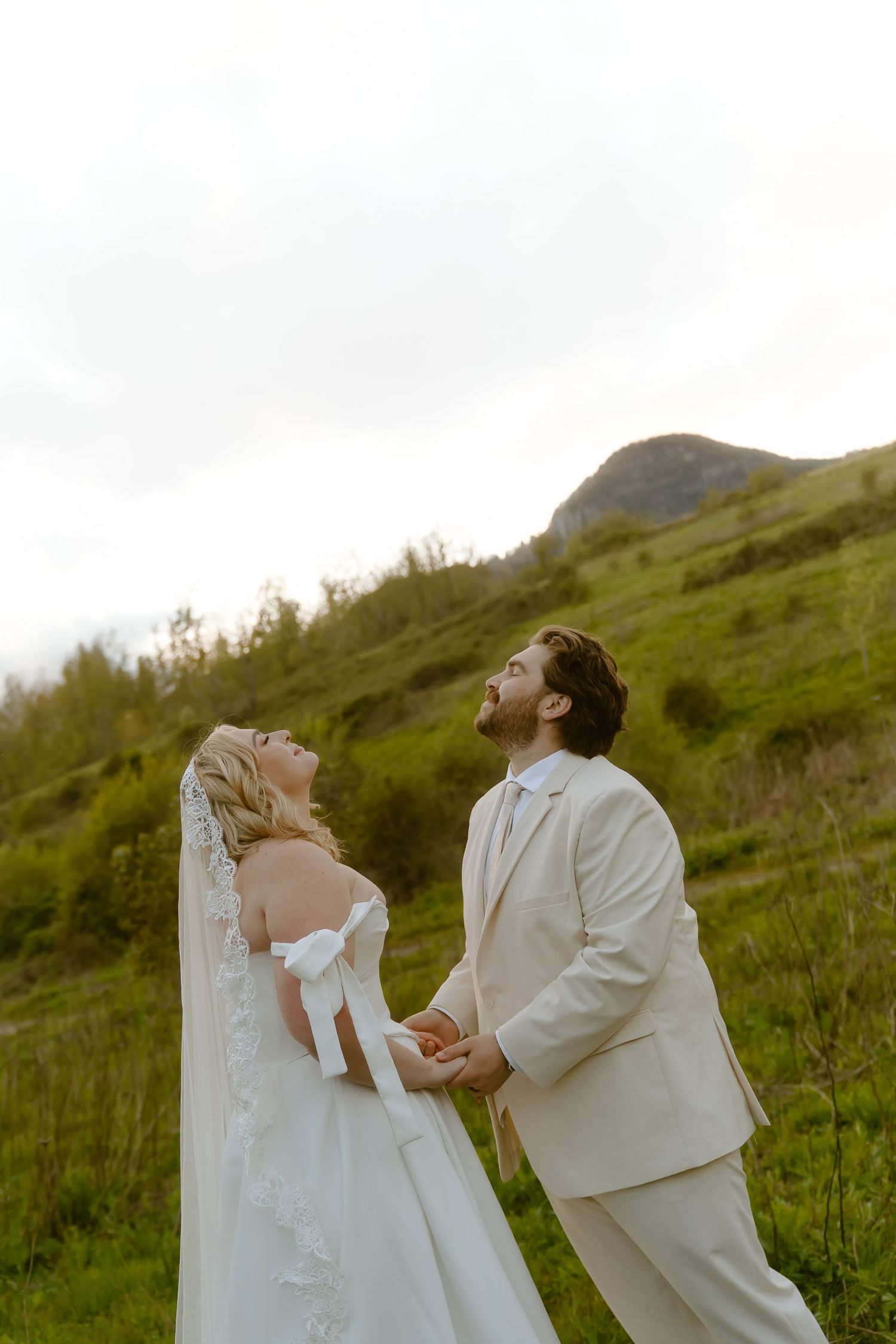 A bride and groom hold hands and laugh joyfully outdoors, with lush green hills and a mountain backdrop.