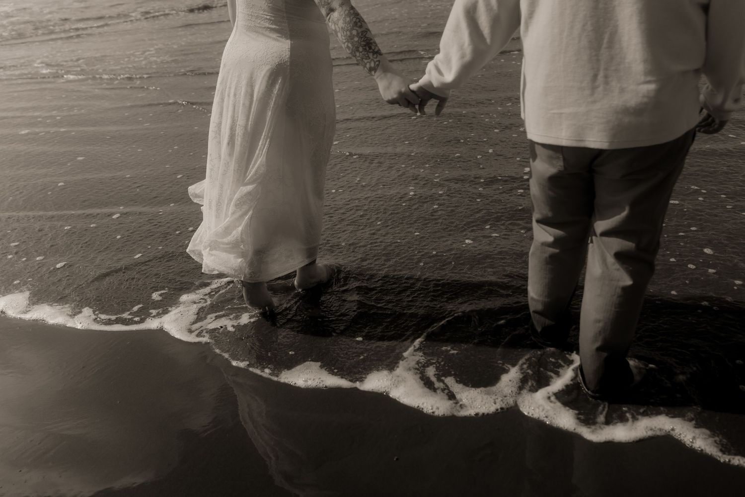A black and white photo shows people standing at the edge of ocean waves on a beach.