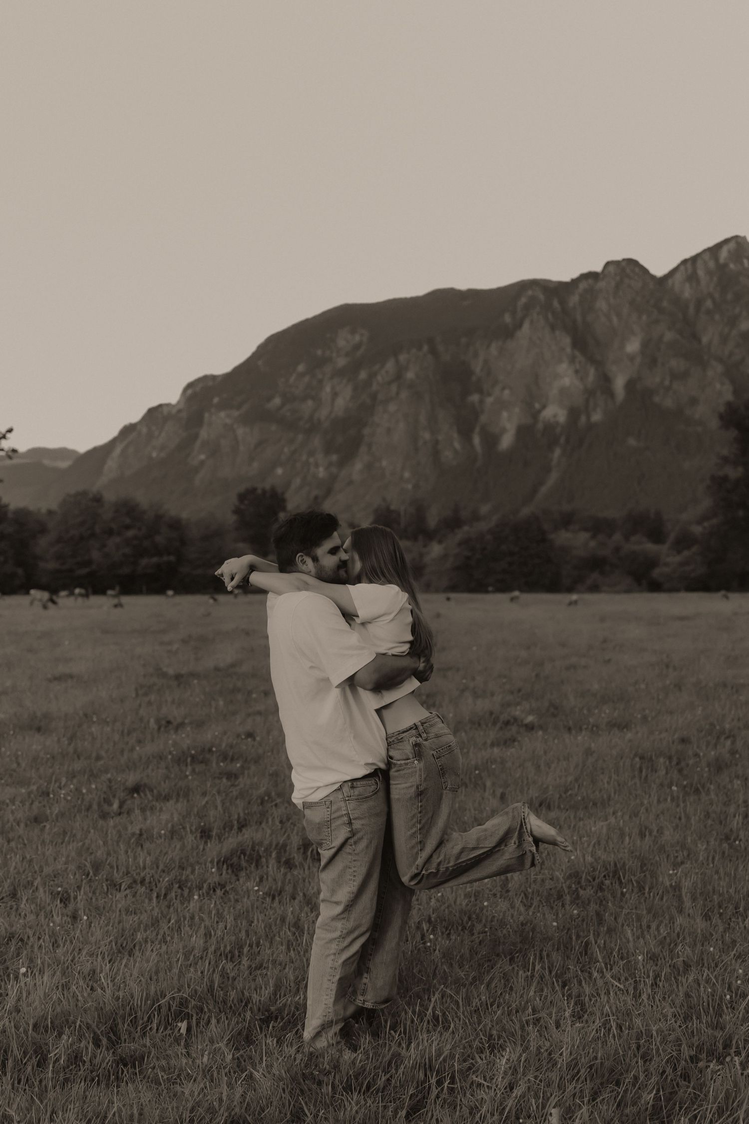 Couple embracing romantically in a field with mountain backdrop in sepia tone photography.