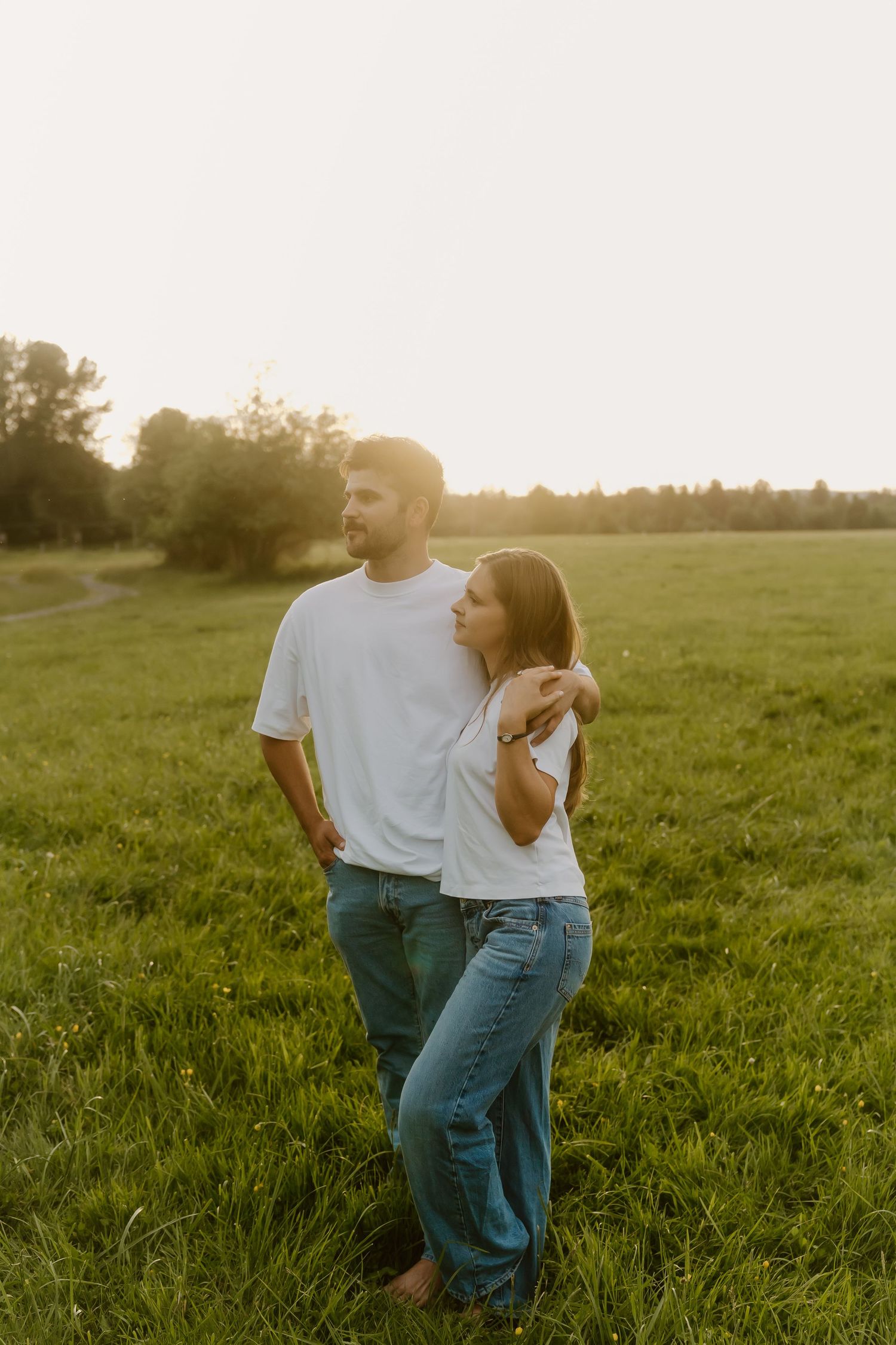 Couple in white t-shirts embracing in a sunlit green field at golden hour.