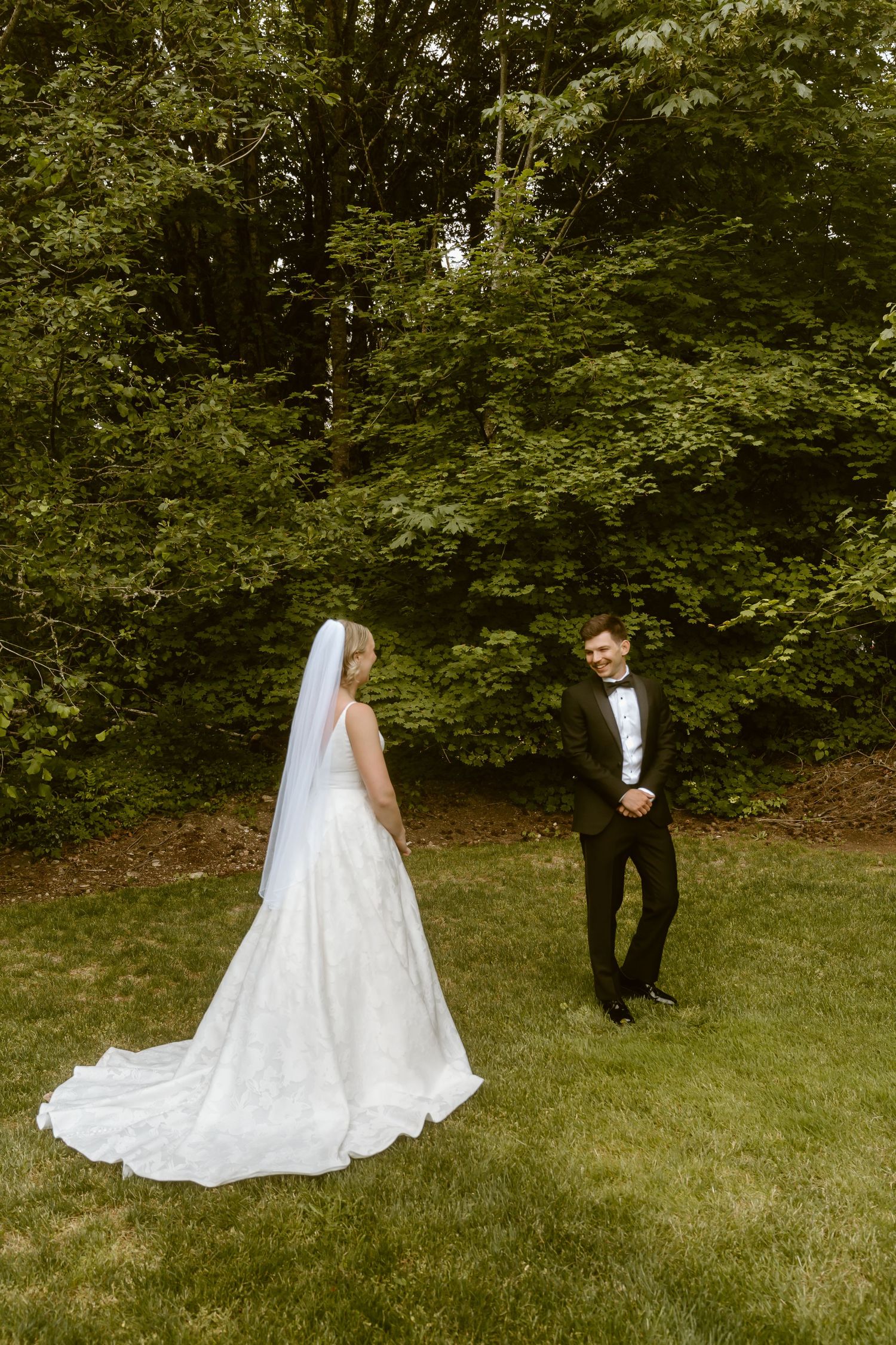 Bride in elegant white gown with long veil faces smiling groom in black tuxedo on garden lawn.